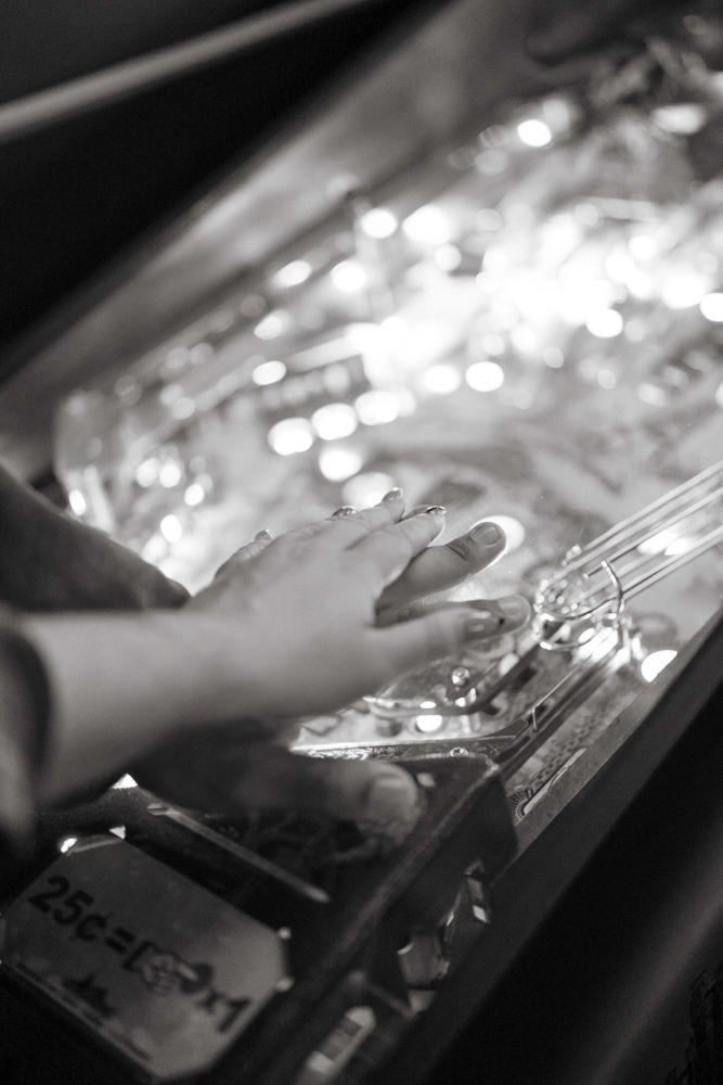 Black and white close-up of hands playing a pinball machine with bright flashing lights.