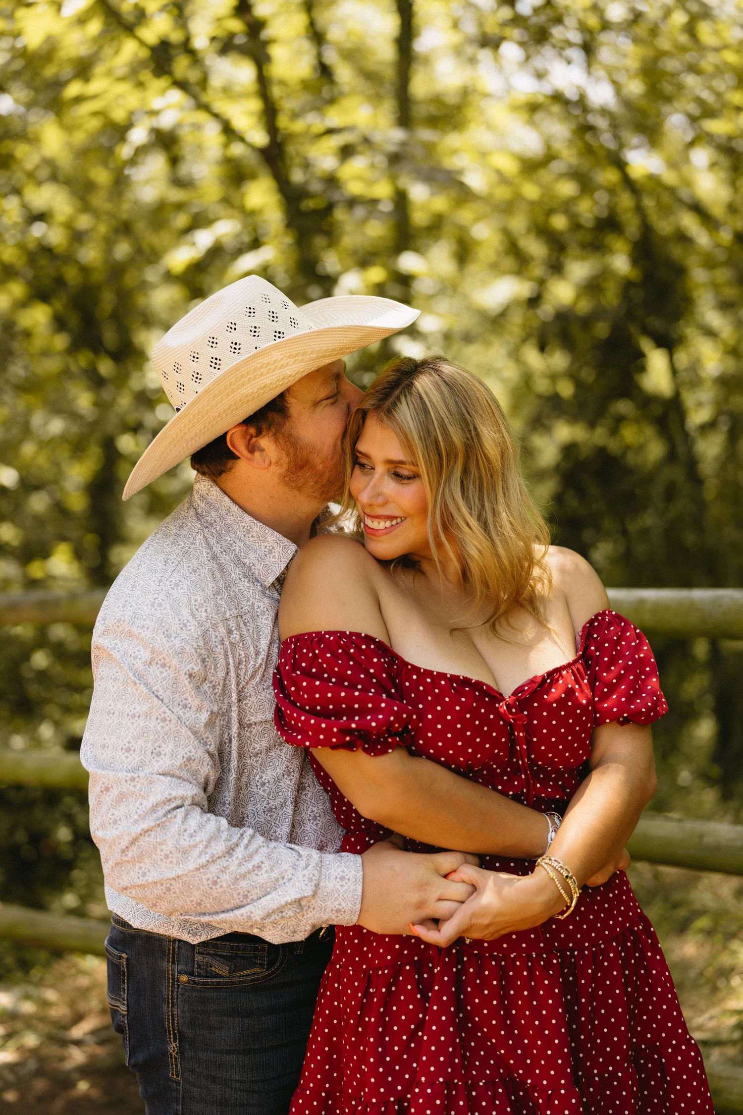 A couple in western attire embraces outdoors, man in cowboy hat kissing woman in red polka dot dress.