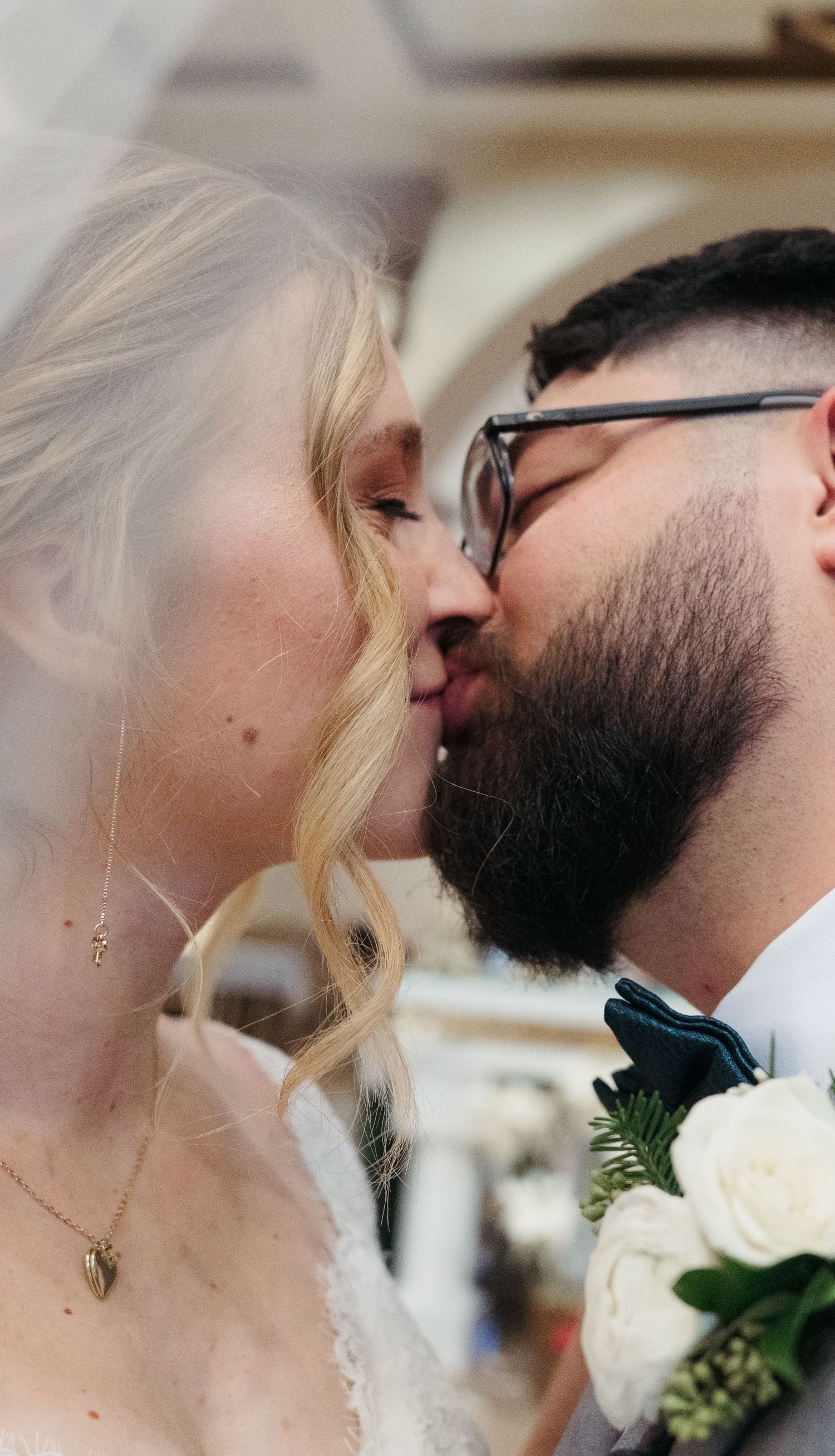 Close-up of bride and bearded groom sharing first kiss, bride wearing gold heart necklace and white veil.