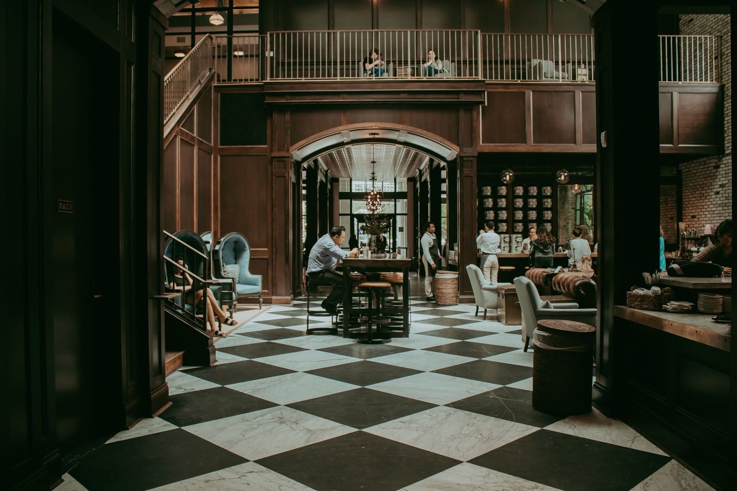 Elegant hotel lobby with black and white checkered floors, wood paneling, arched doorway, and guests seated at tables.