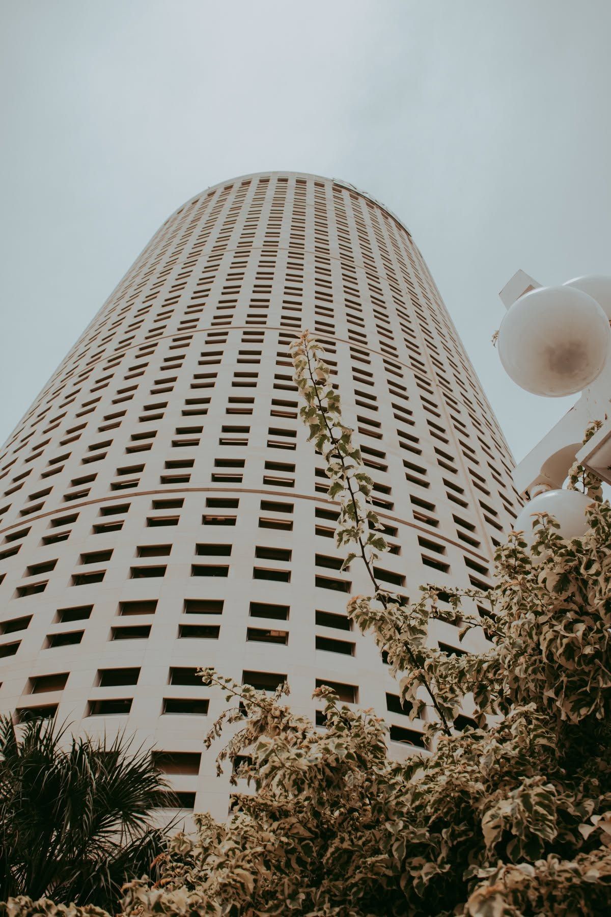Low-angle view of a tall cylindrical skyscraper with grid windows, surrounded by trees and a street lamp.