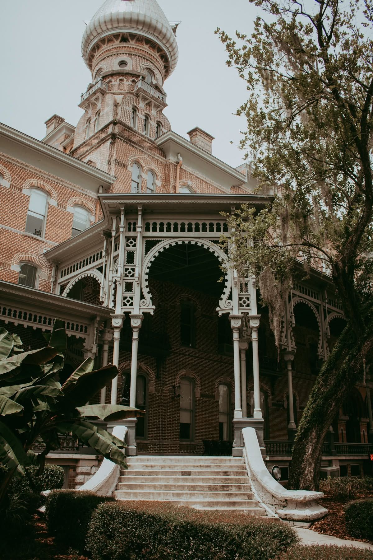 Historic Victorian brick building with ornate white porch, decorative columns, steps, and a domed tower surrounded by lush trees.