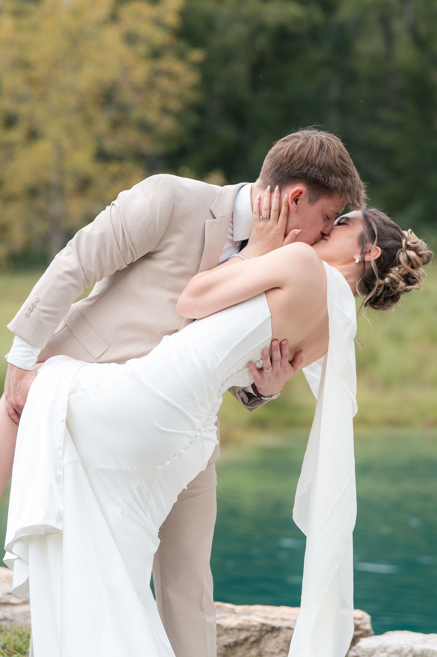 Groom in tan suit dips bride in white wedding dress for romantic kiss near turquoise pond.