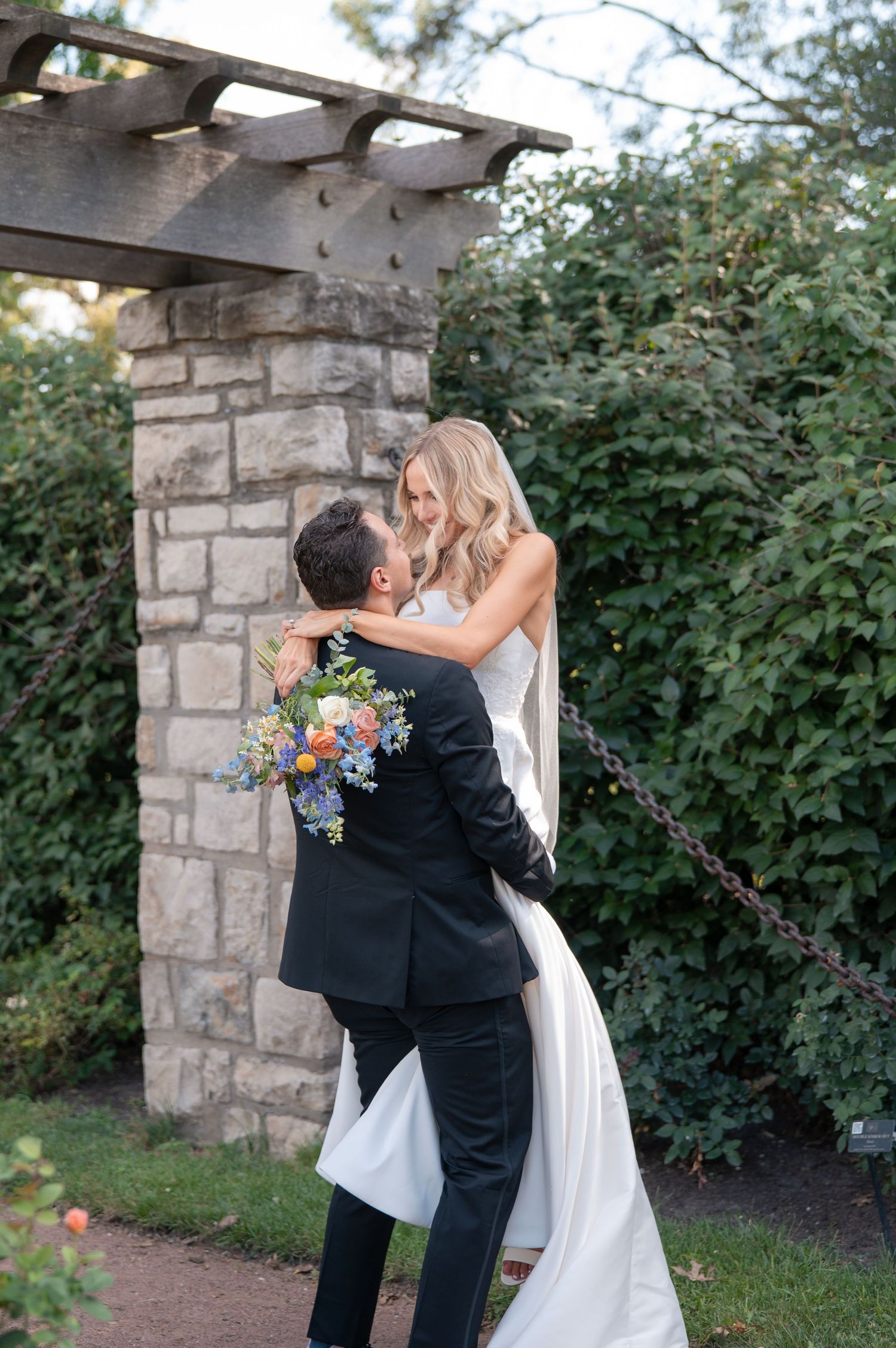 Groom lifts smiling bride holding colorful bouquet under stone pergola surrounded by greenery.