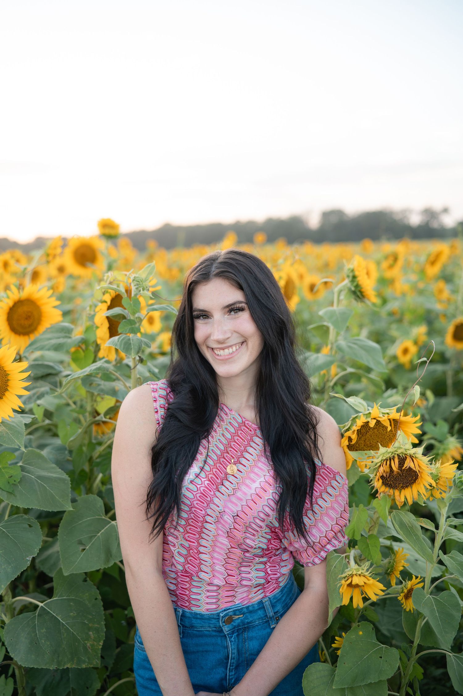 Brunette teen girl in pink patterned top smiling in a sunflower field during golden hour.