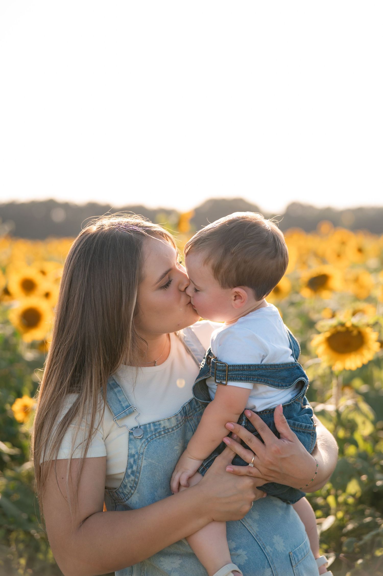 Mother kissing toddler boy in denim overalls in a sunny sunflower field at golden hour.