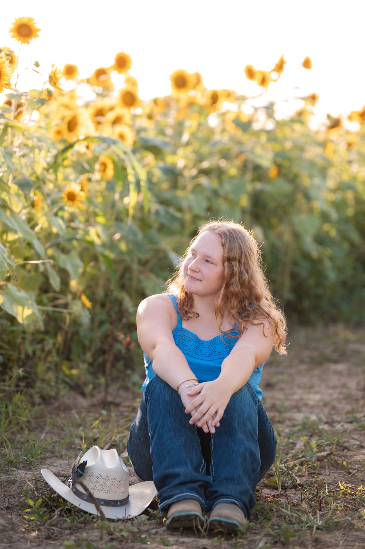 Young woman in blue tank top and jeans sitting in sunflower field with cowboy hat beside her.