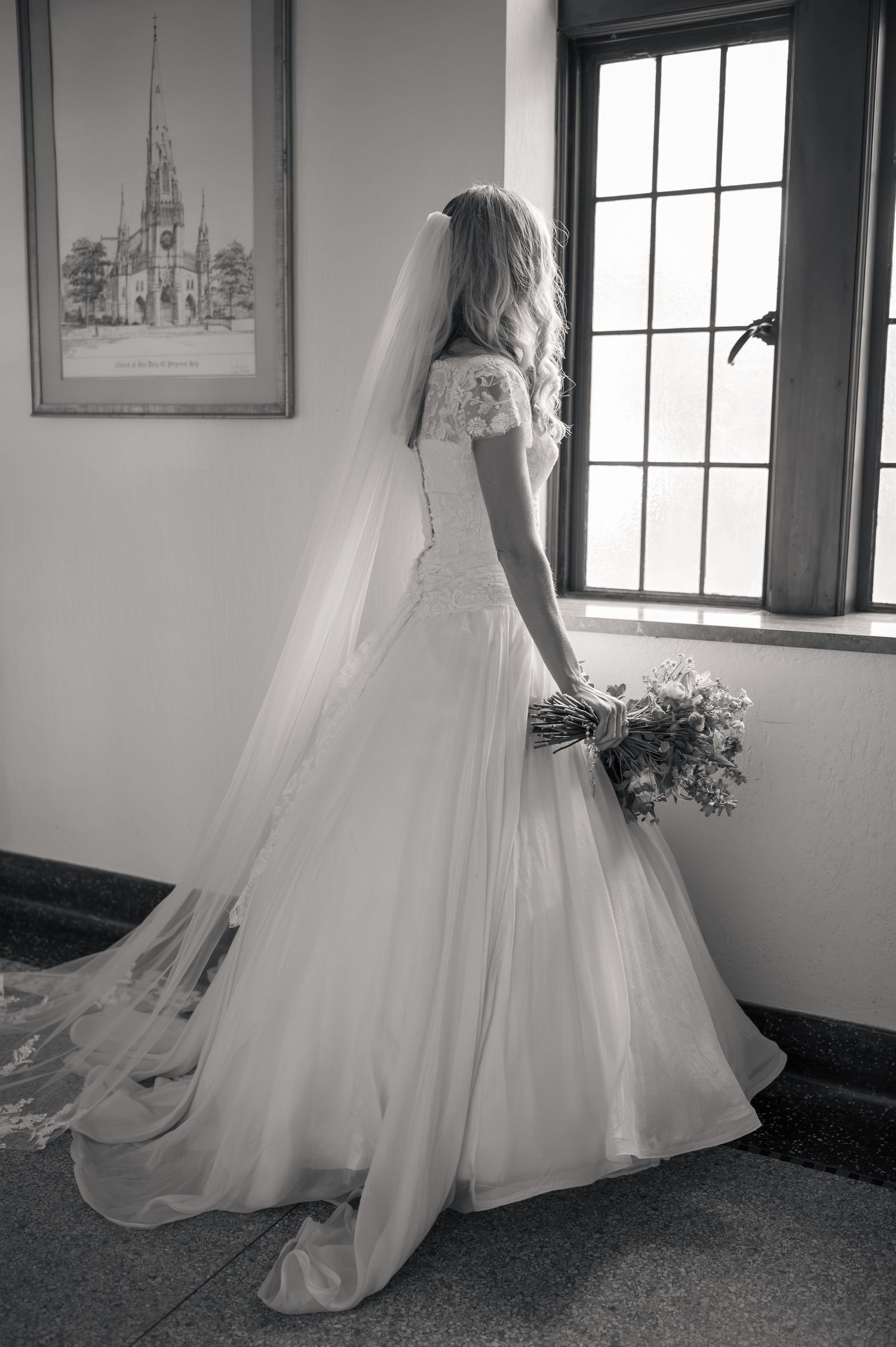 Bride in off-shoulder lace gown with cathedral veil gazes out a church window holding a bouquet.