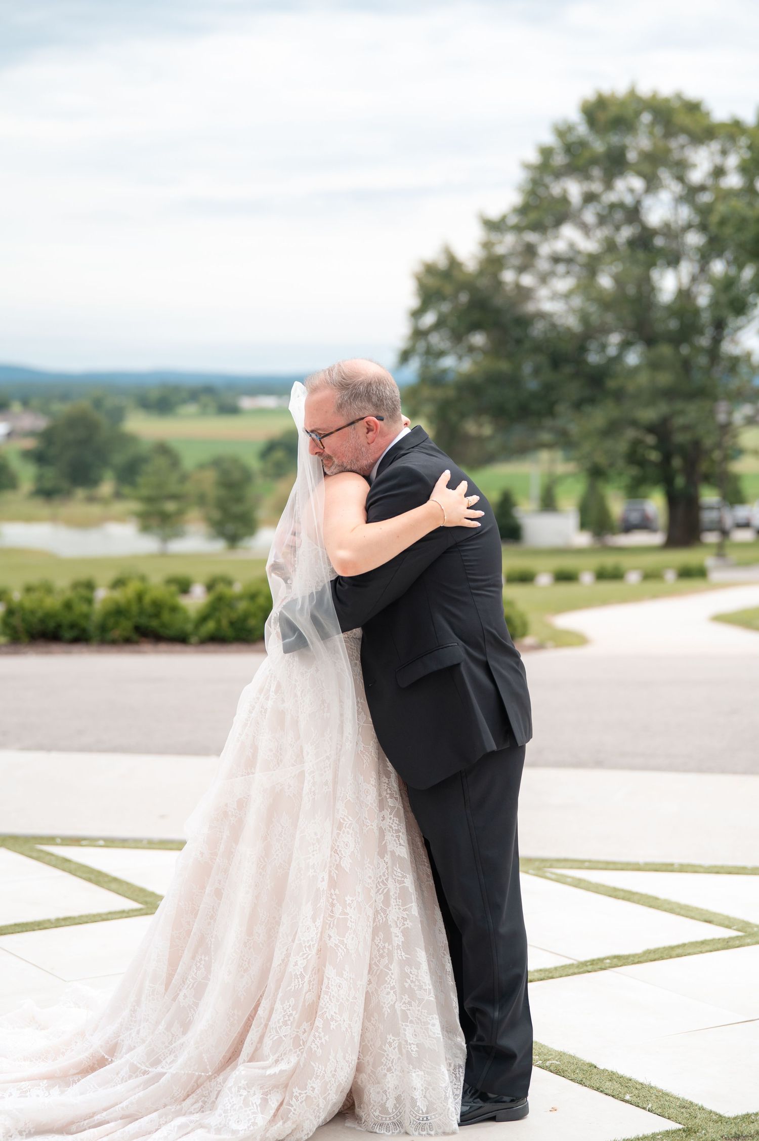 Bride in lace gown sharing emotional first look hug with father outdoors.