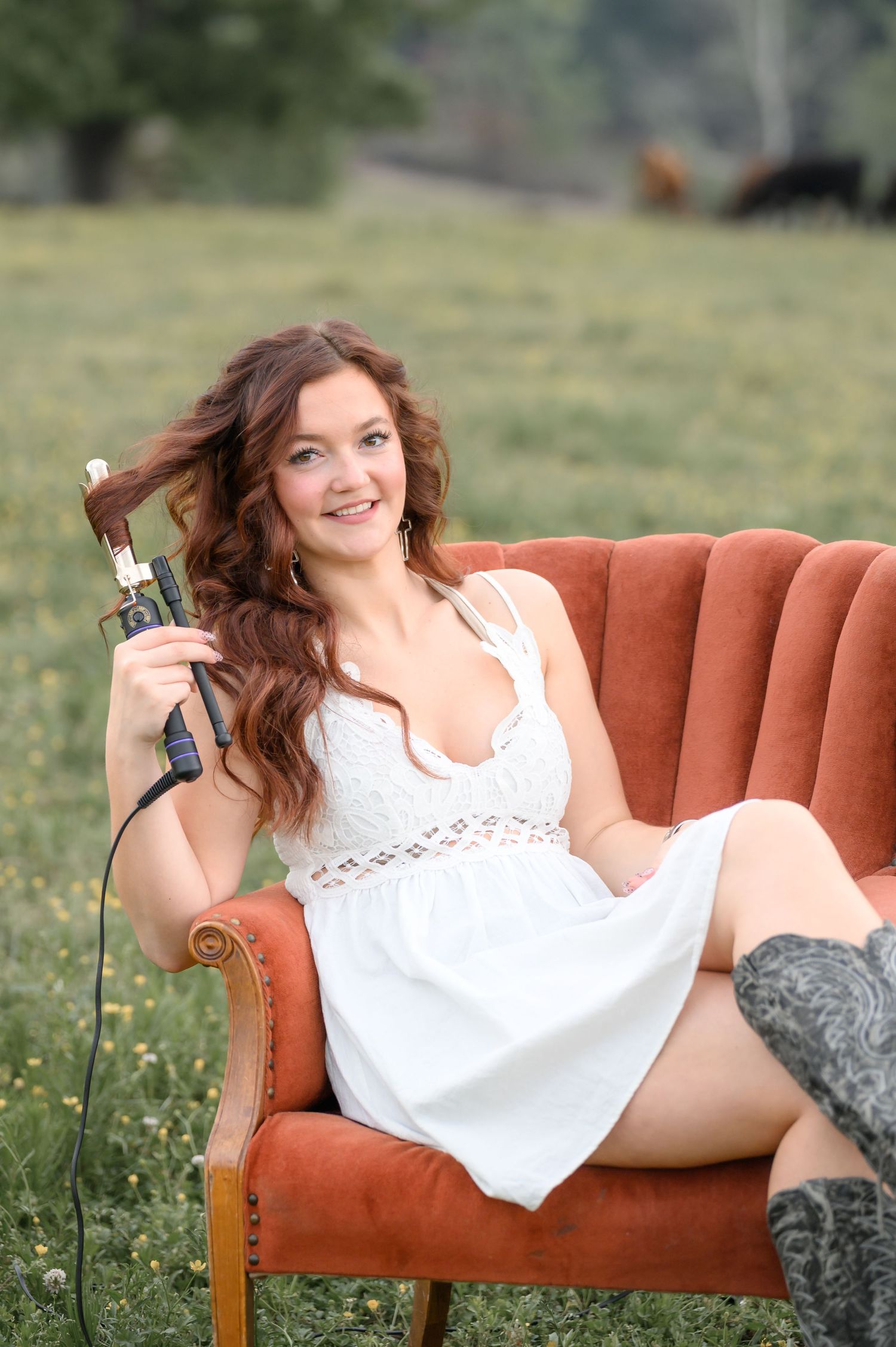Young woman in white dress curling her hair with a curling iron while sitting in a red velvet chair outdoors.