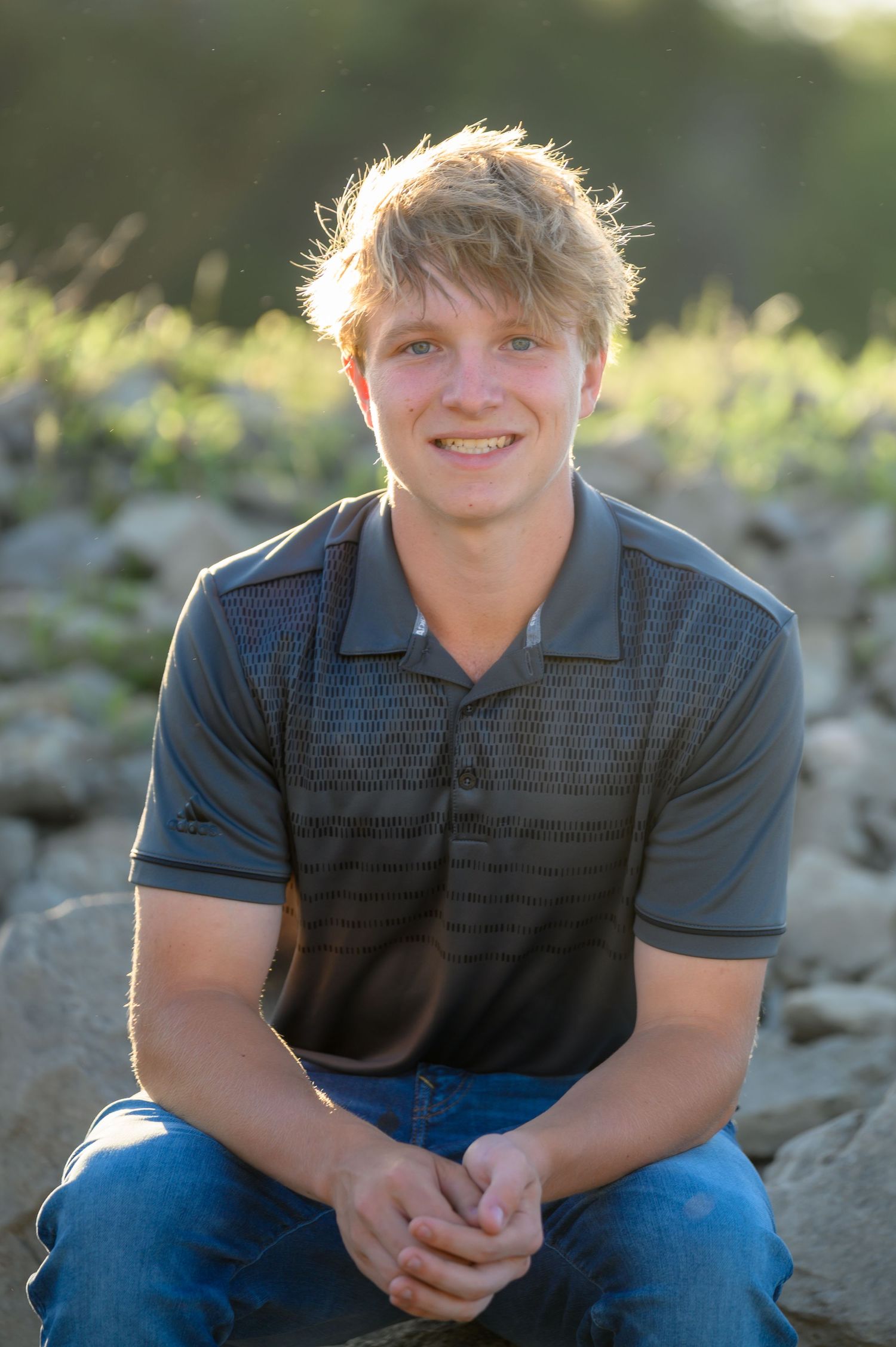 Smiling blonde teenage boy in a gray polo shirt sitting on rocks in golden hour sunlight.