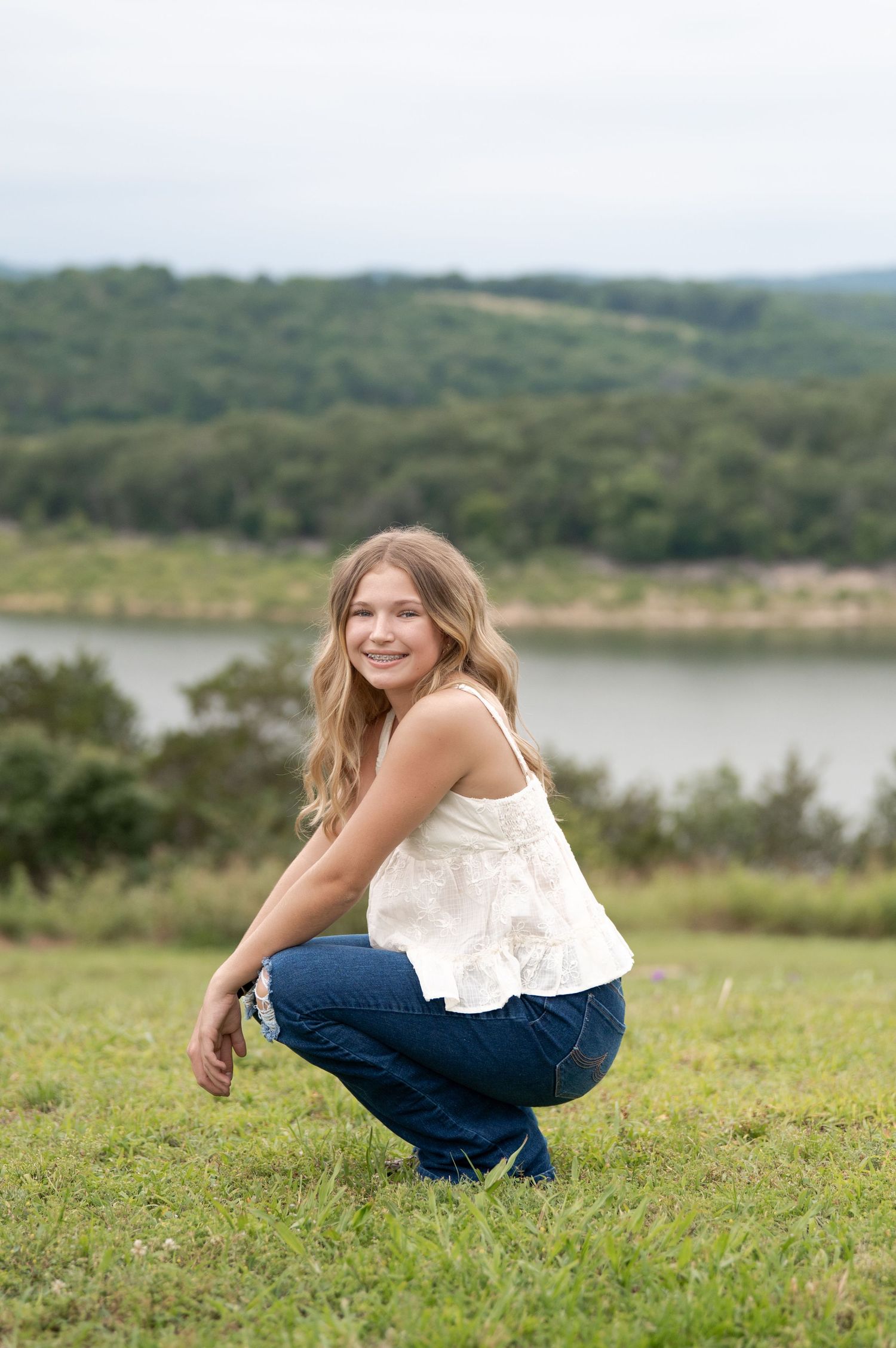 Teenage girl crouching on grass in white top and jeans, smiling with a lake backdrop.
