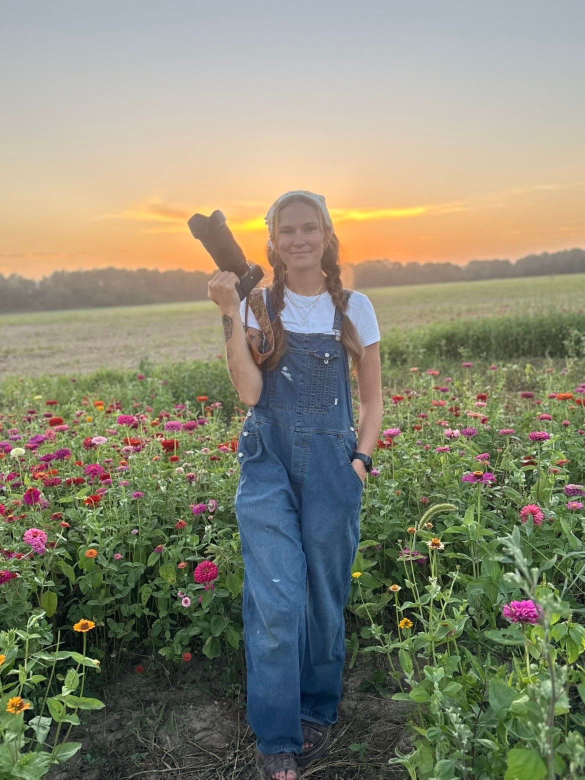 Young woman in denim overalls holding boots in a colorful zinnia flower field at golden sunset.