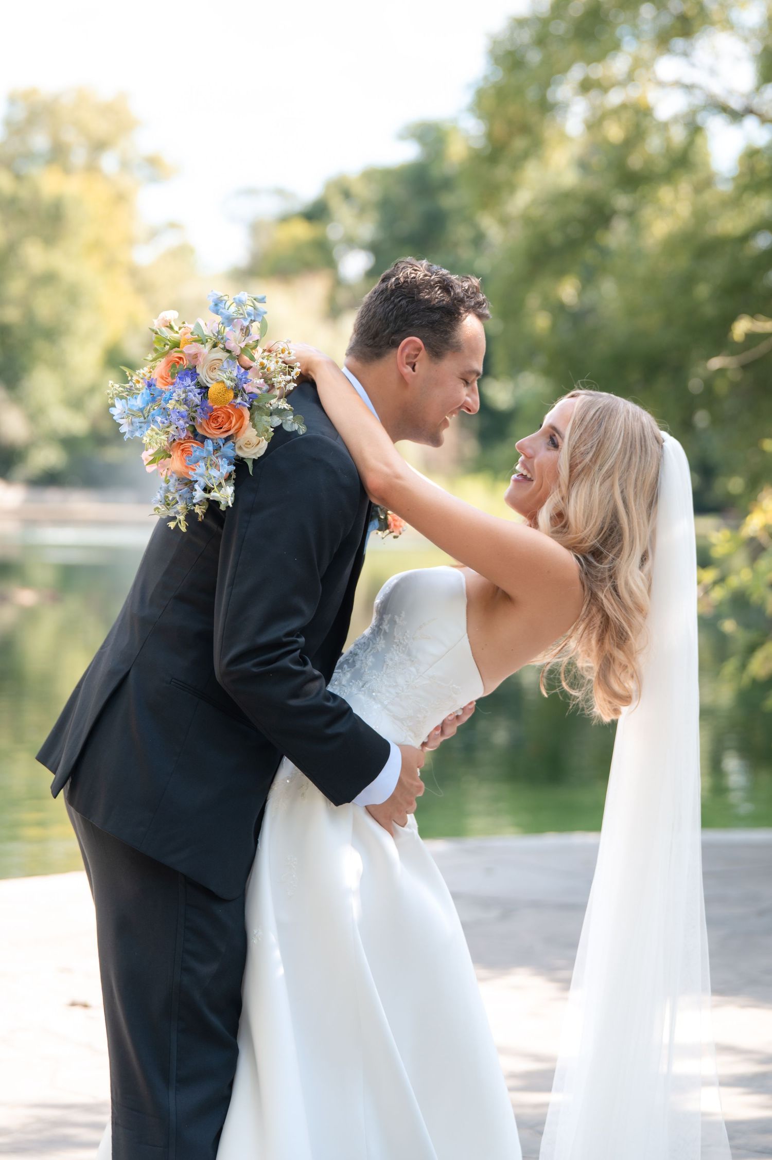 Bride and groom embrace lakeside, bride holding vibrant blue and orange wedding bouquet.