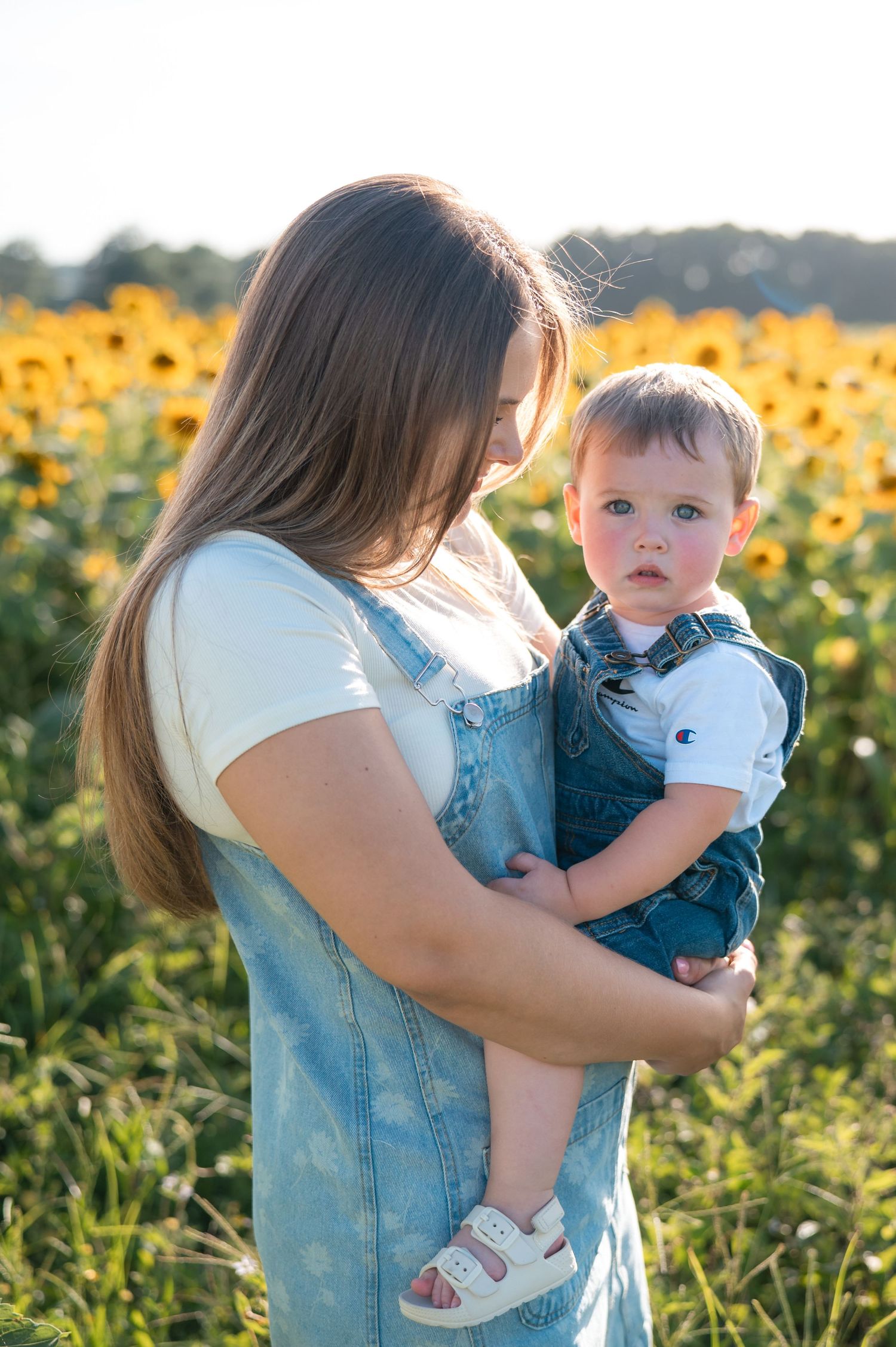 Mother in denim overalls holding curious toddler boy in sunflower field at golden hour.