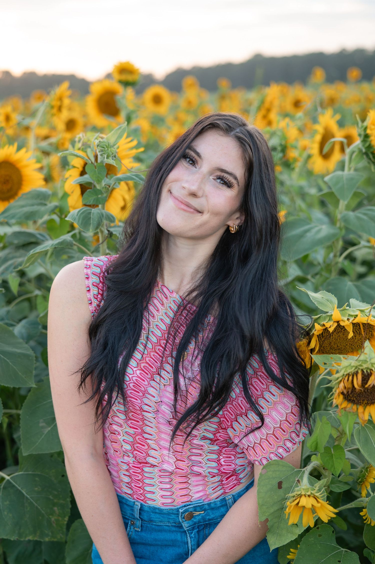 Dark-haired young woman in pink top smiling softly among sunflowers during golden hour.