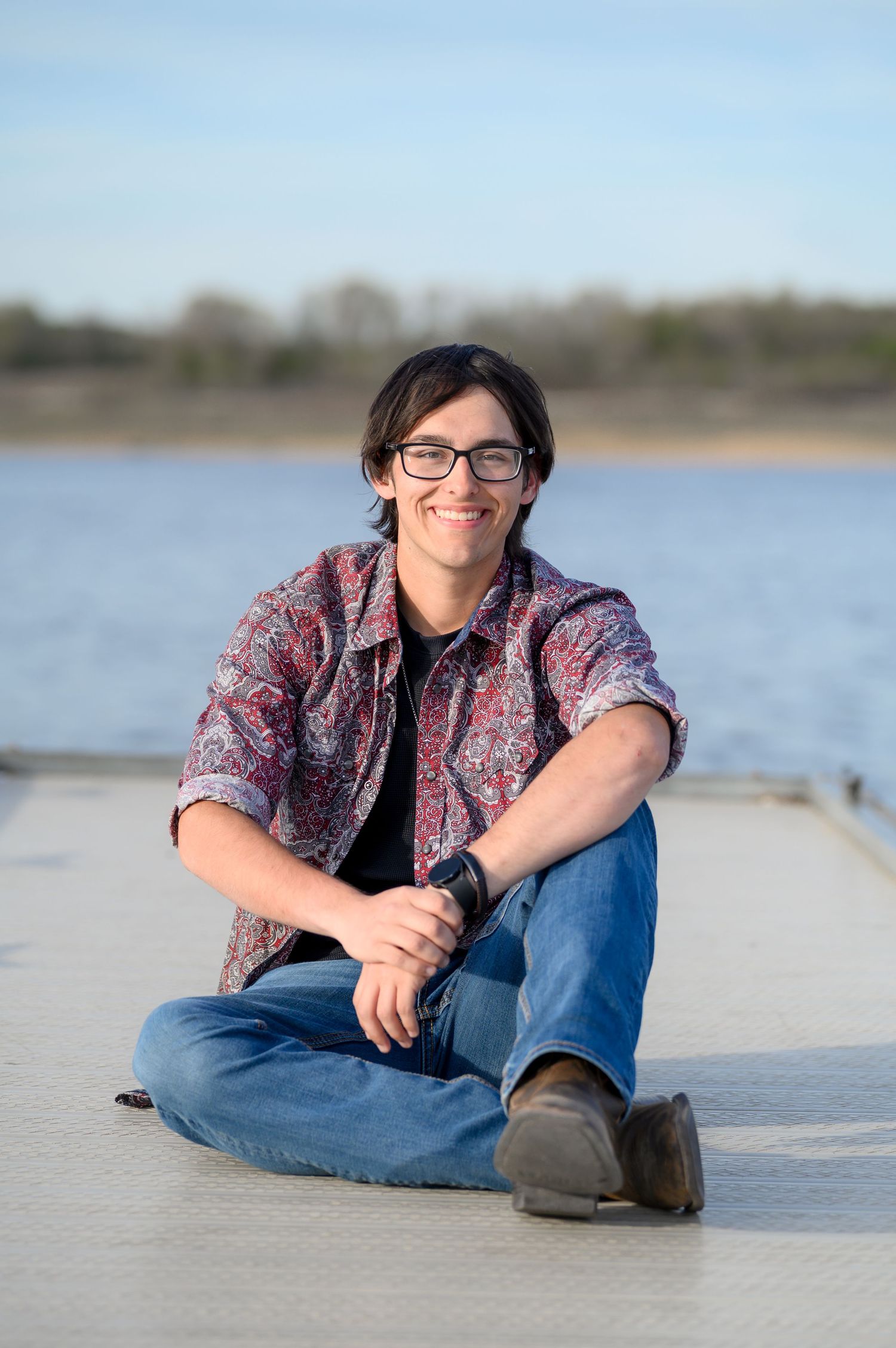 Smiling young person with glasses sitting cross-legged on a dock beside a calm lake.