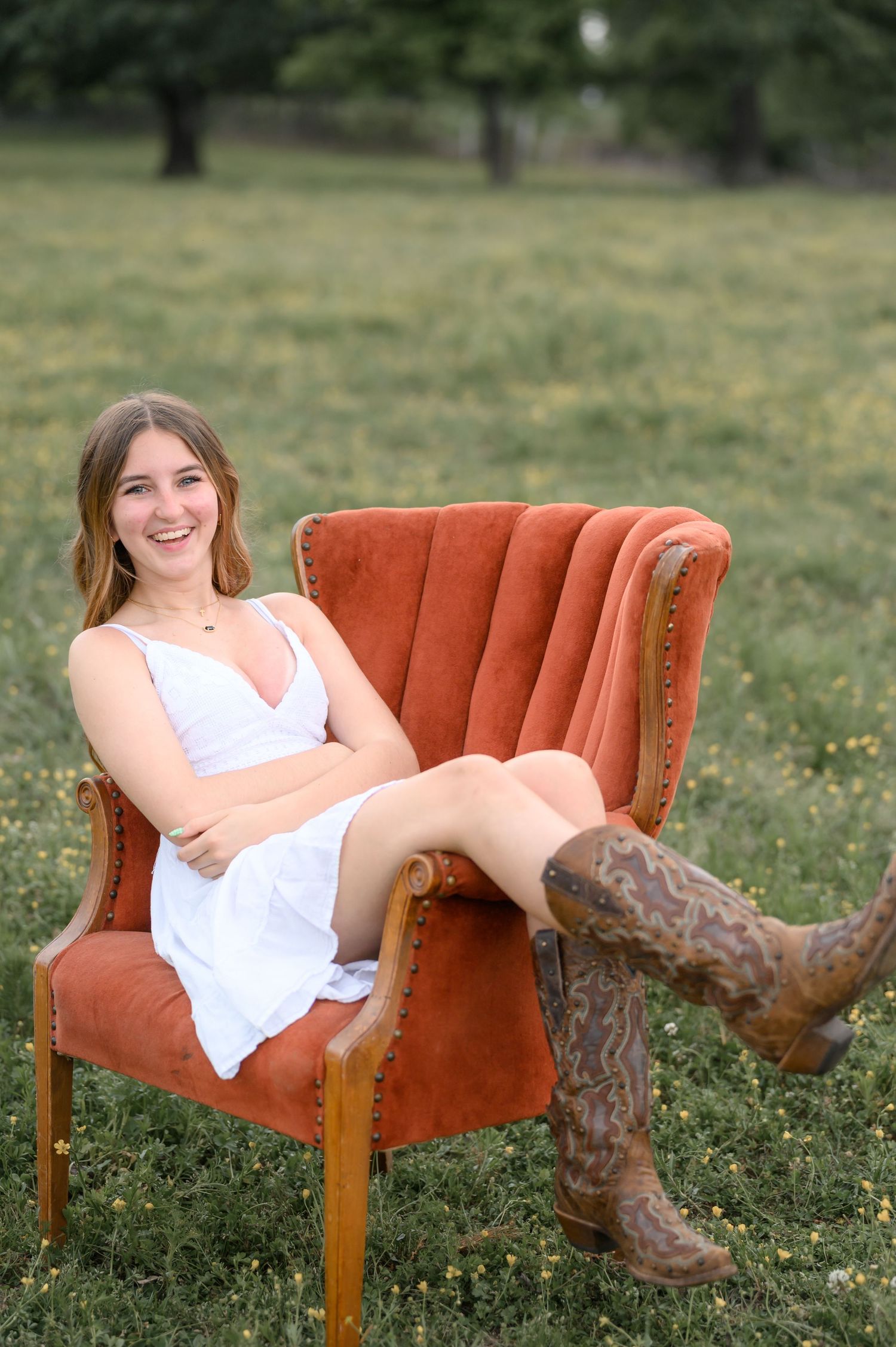 Smiling young woman in white dress and cowboy boots relaxing in an orange velvet chair in a green field.