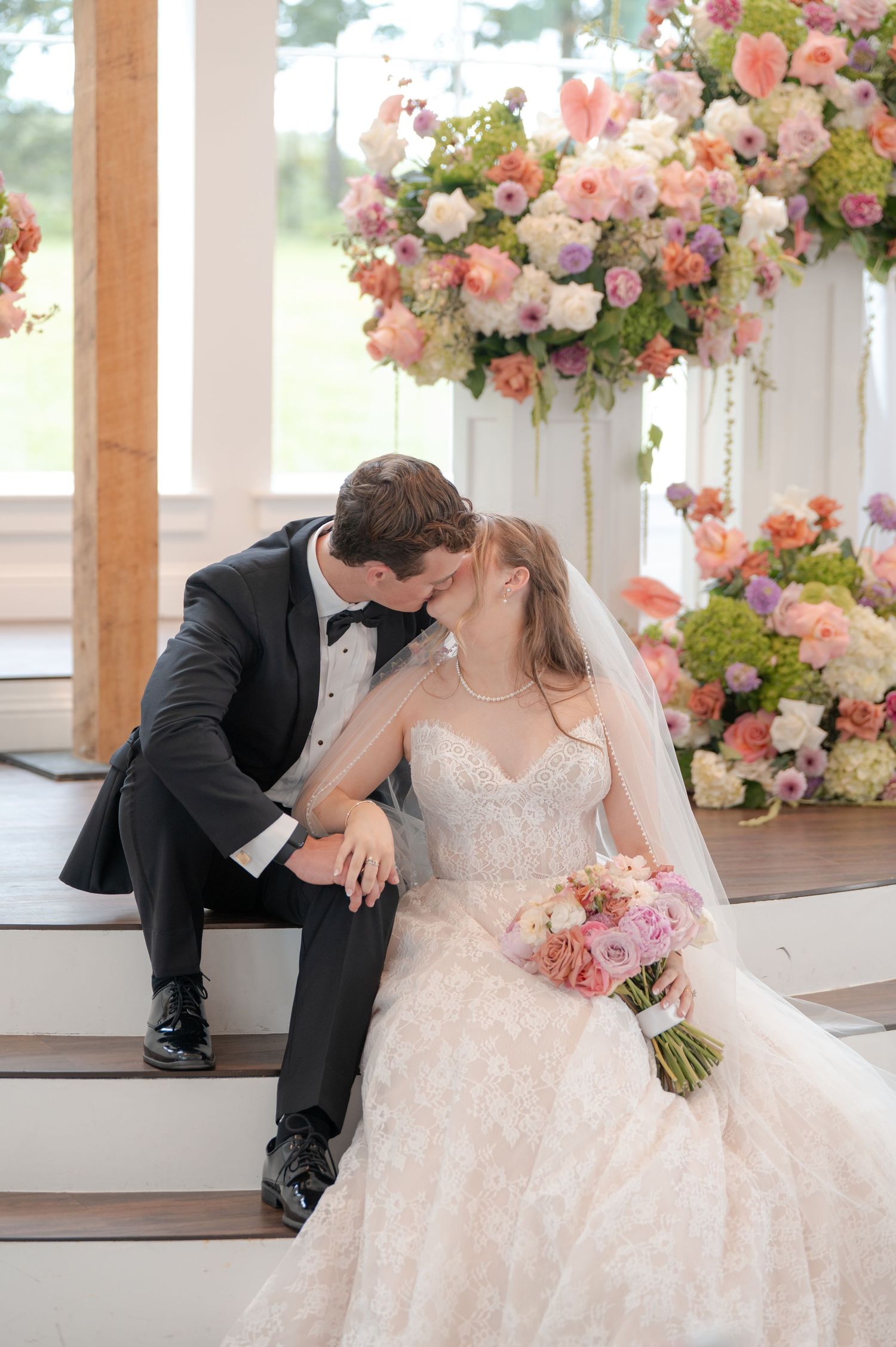 Bride and groom share tender kiss on steps surrounded by lush pink and coral floral arrangements.