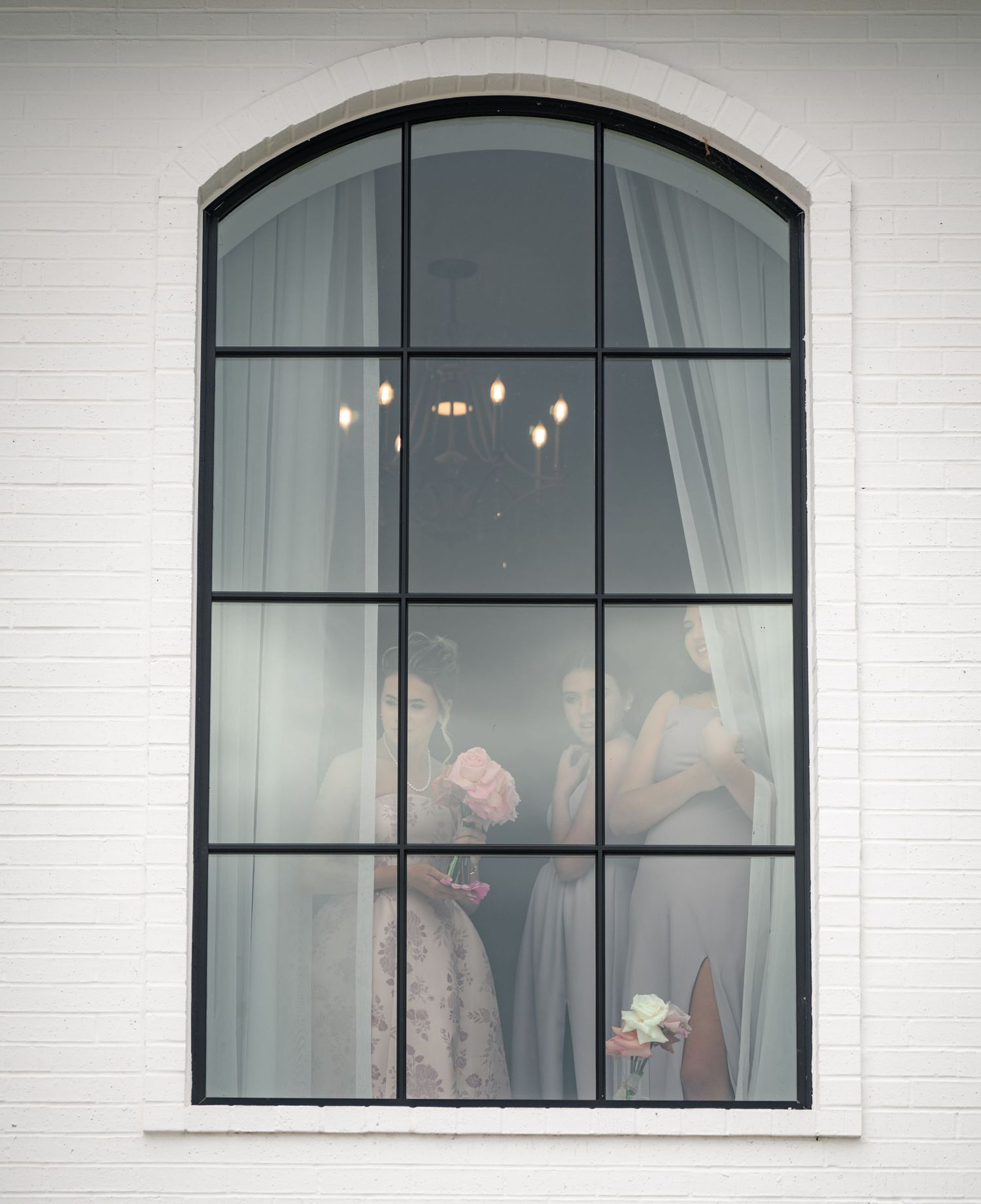 Bridesmaids visible through arched window of white wedding venue holding flowers.