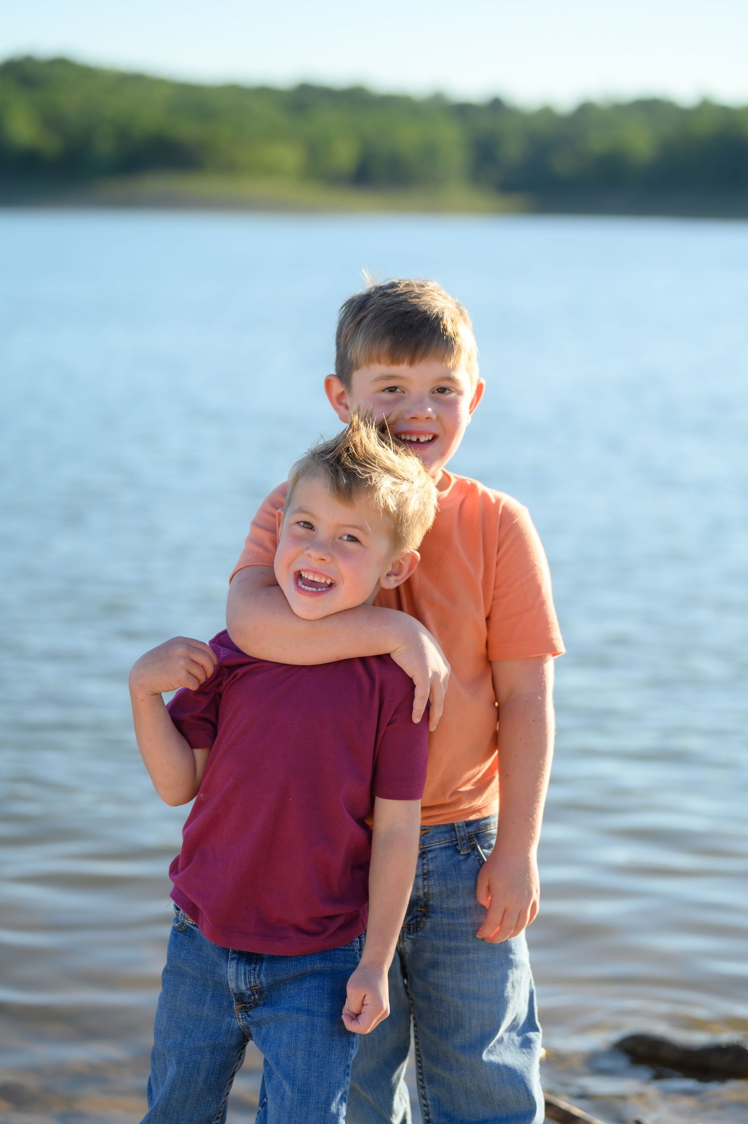Two smiling boys hugging near a lake shore on a sunny day.