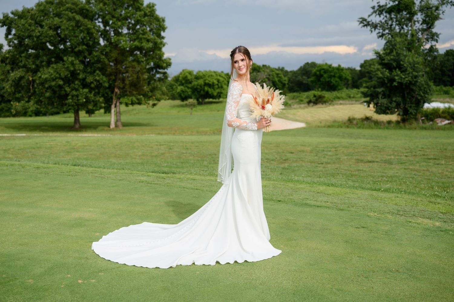 Bride in fitted white lace long-sleeve gown with cathedral train holding bouquet on golf course.