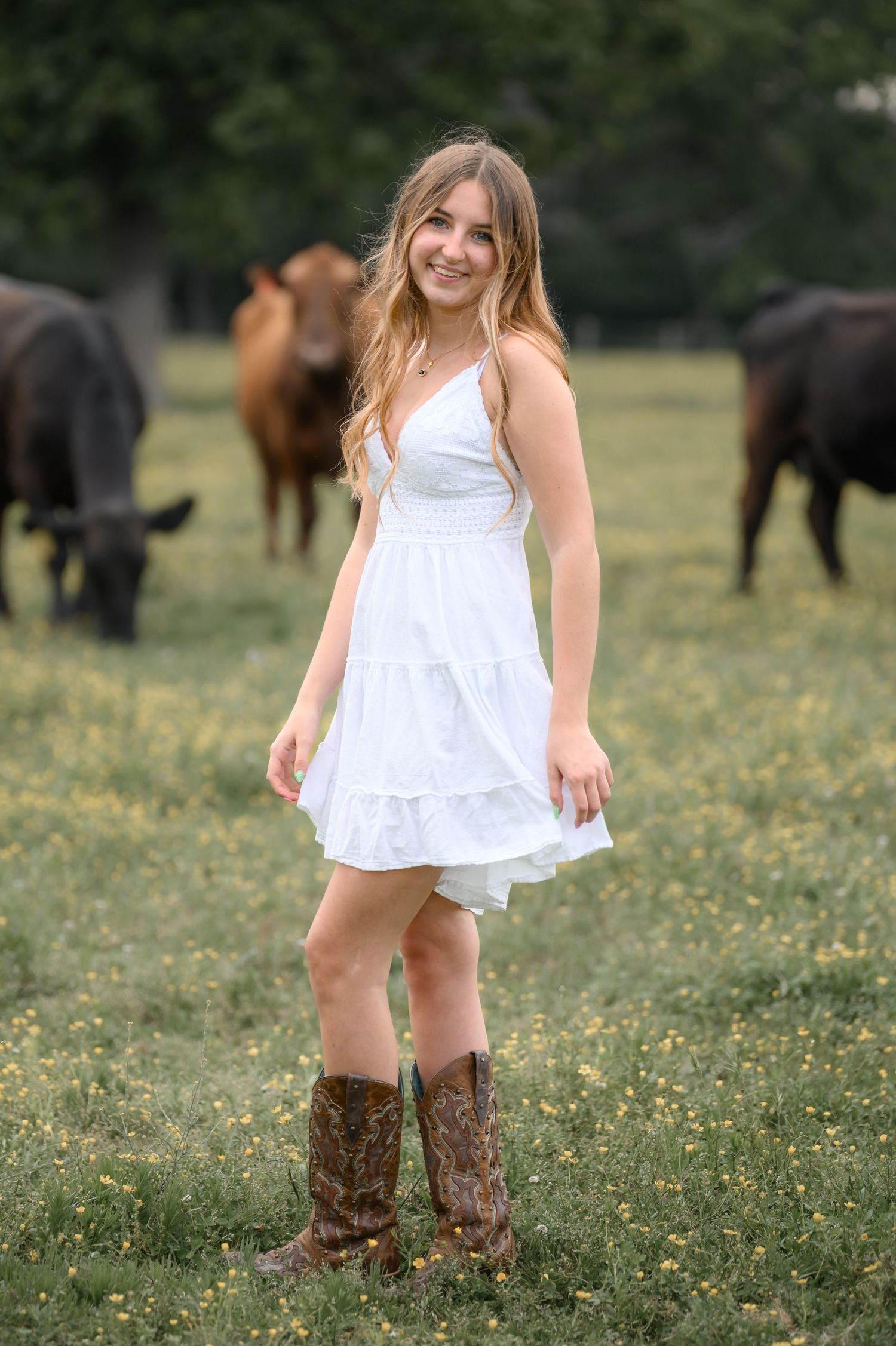 Young woman in white dress and cowboy boots smiling in a pasture with cattle grazing.