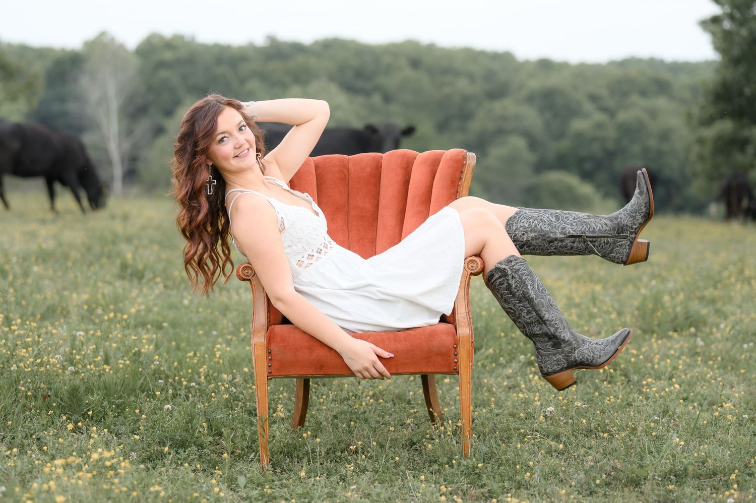 Young woman in white dress and tall boots posing elegantly in a velvet chair in a pasture.