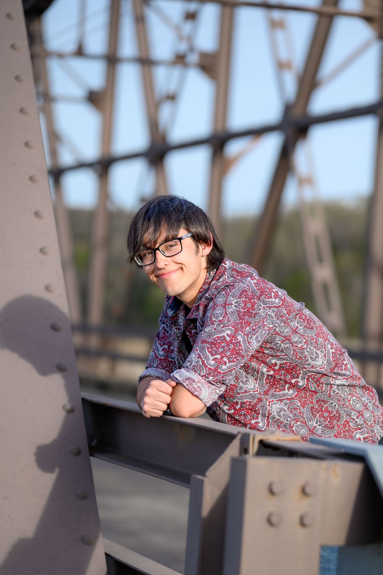 Young person with glasses in a paisley shirt leaning on a metal bridge structure outdoors.