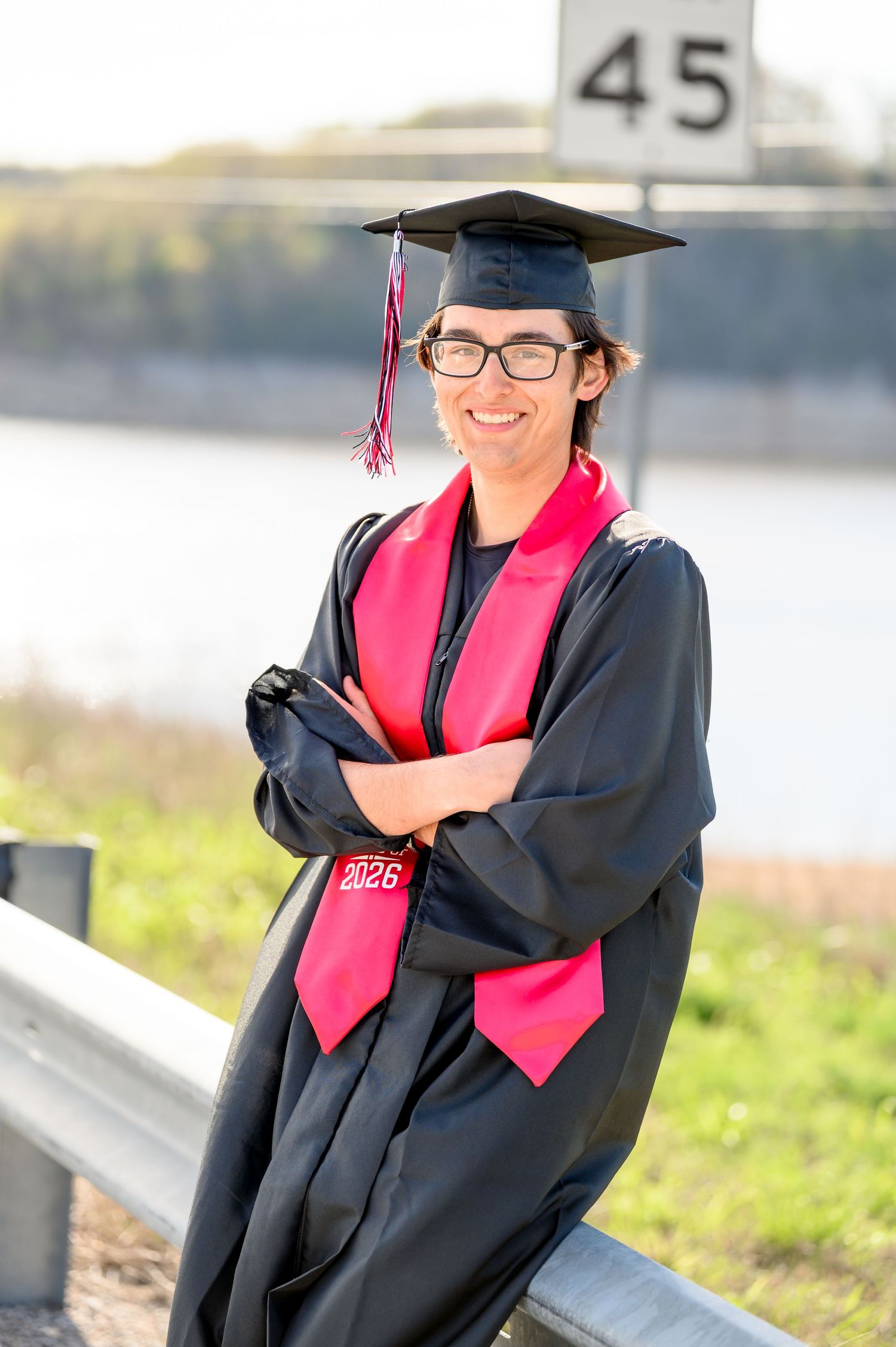 Smiling graduate in black cap and gown with red stole posing outdoors near a waterway.