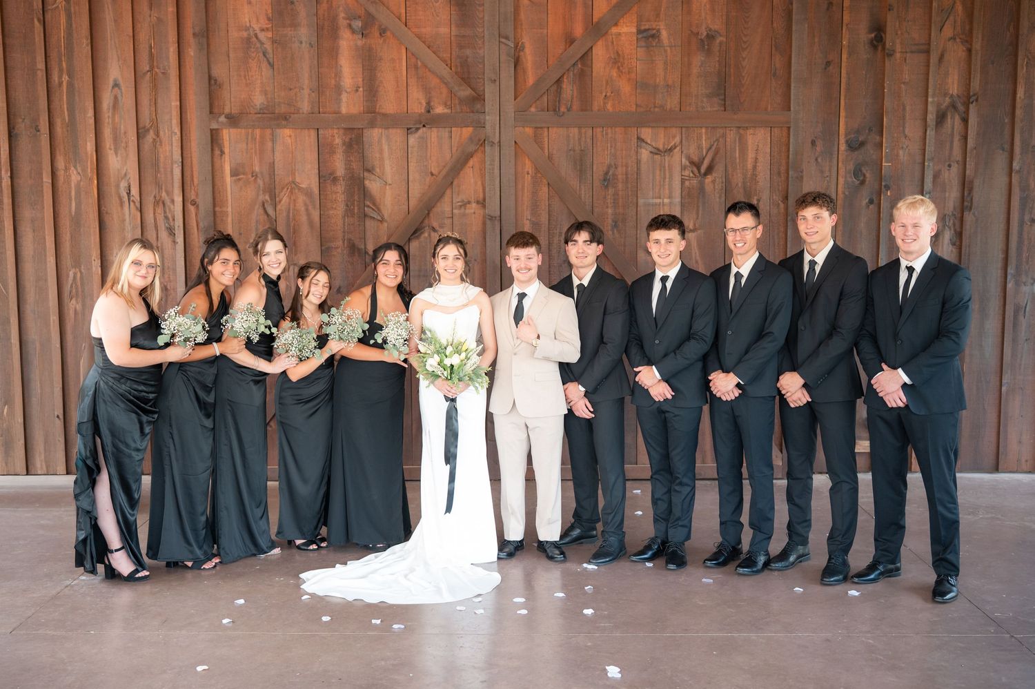 Wedding party poses in a rustic barn, bridesmaids in black gowns and groomsmen in dark suits flanking the bride and groom.