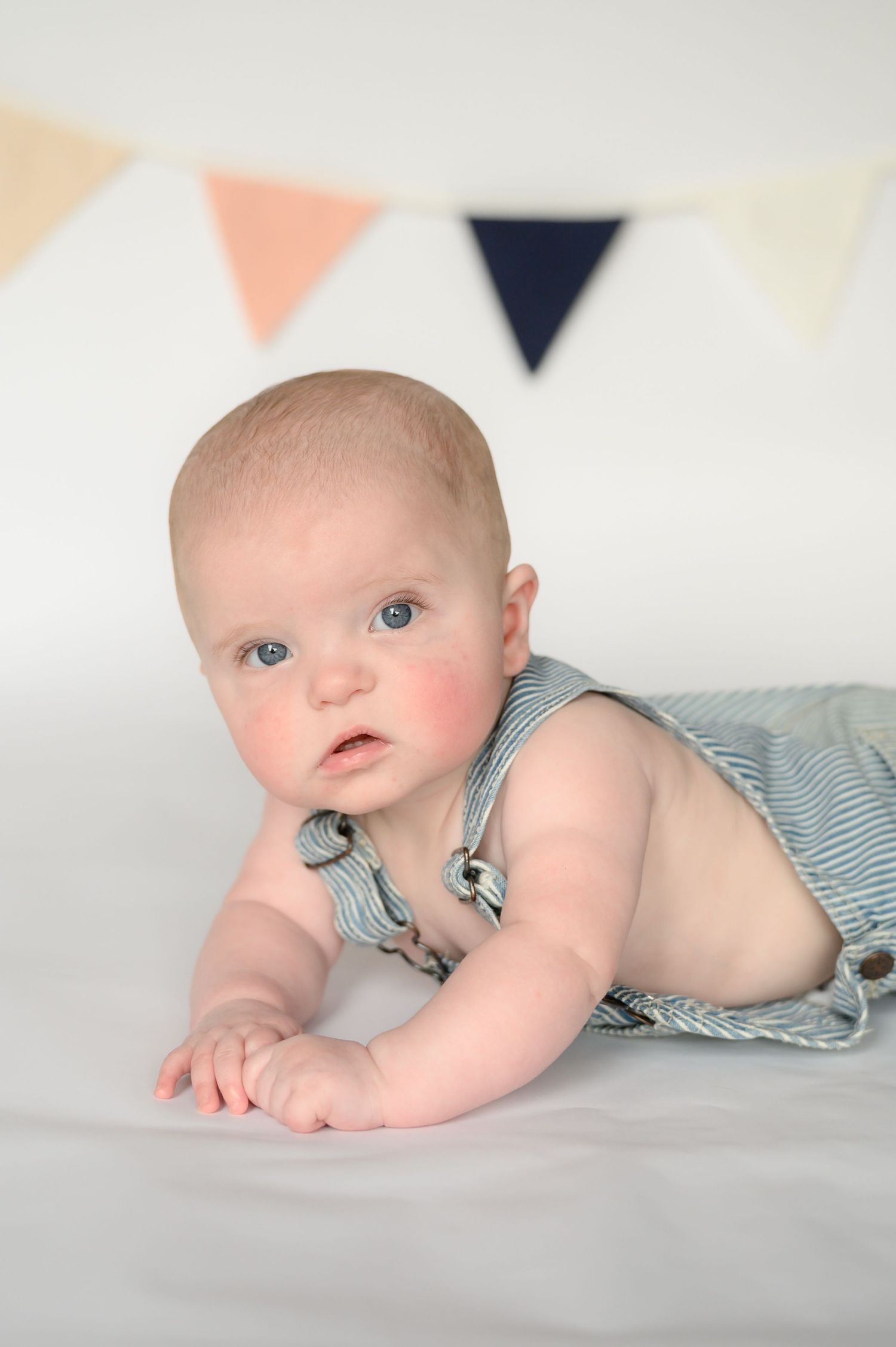 Blue-eyed baby in striped overalls doing tummy time on white surface with colorful pennant banner backdrop.