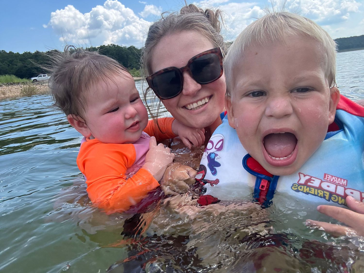 Woman and two toddlers in life jackets enjoying a swim in a lake on a sunny summer day.