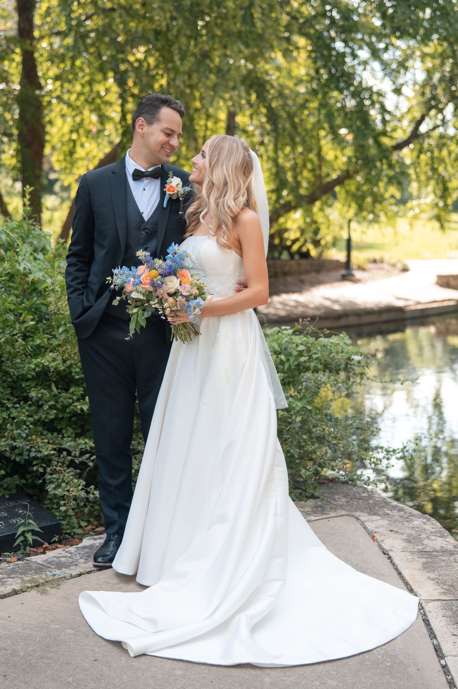 Bride and groom pose by pond in garden, bride holding colorful floral bouquet.