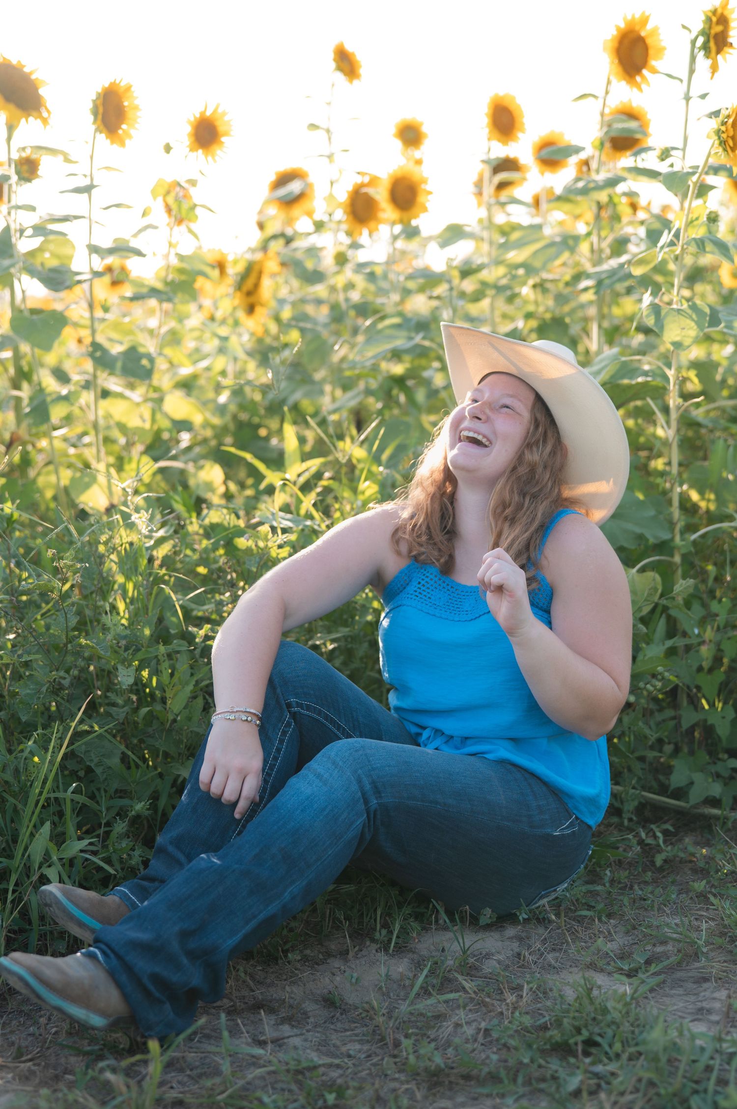 Laughing young woman in blue top and cowboy hat sitting in sunflower field at golden hour.