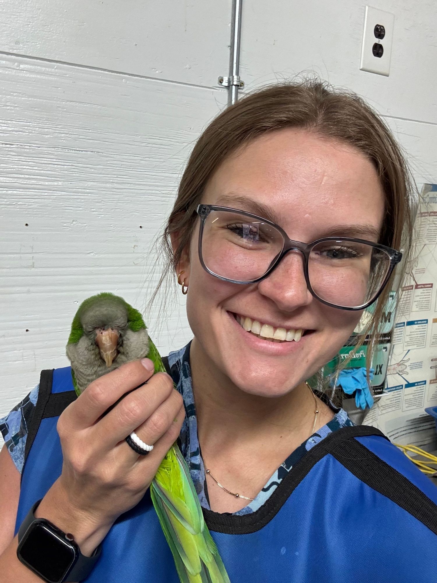 Smiling woman with glasses holds small green parrot while wearing blue scrubs in indoor setting.