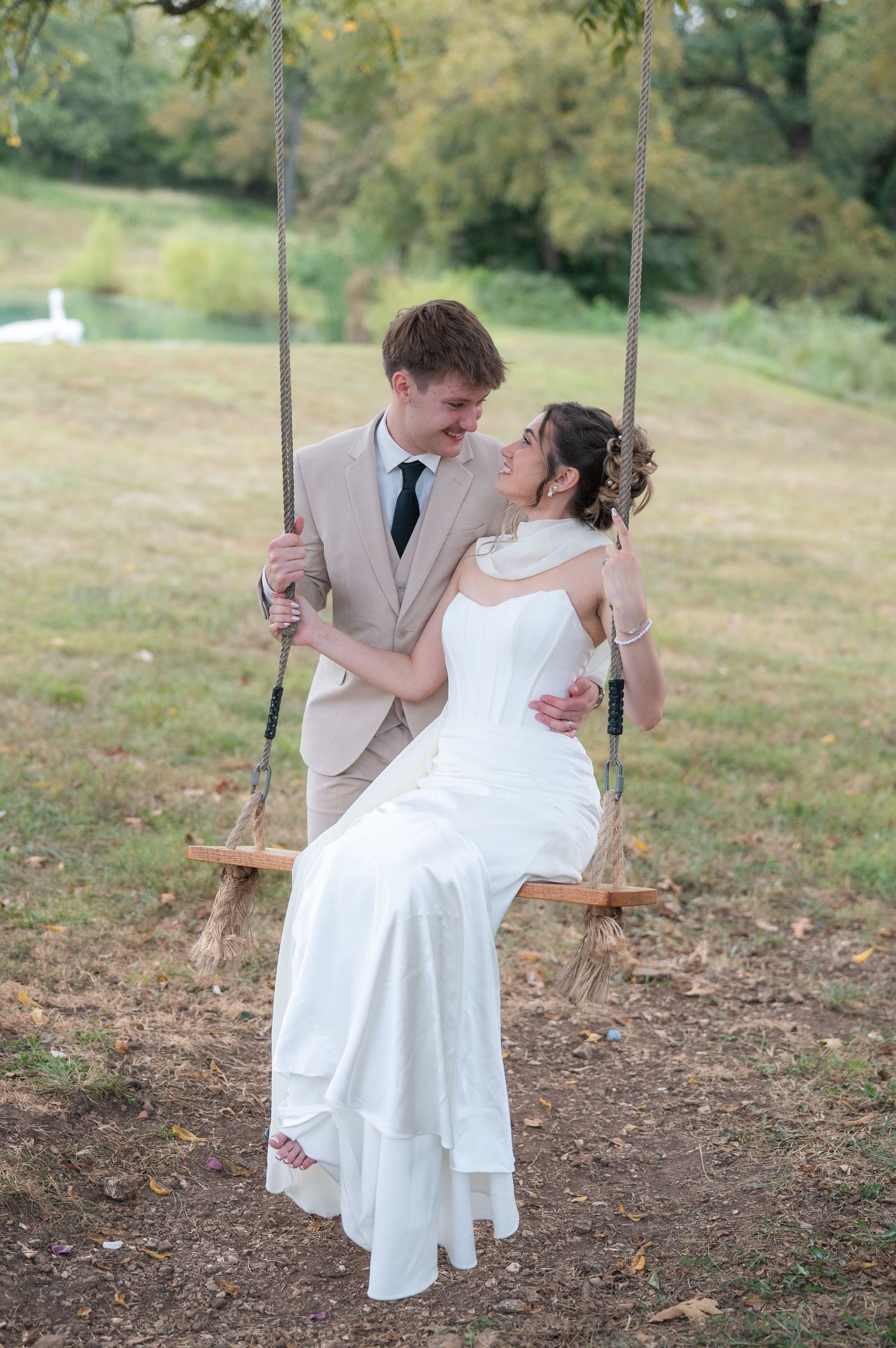 Bride in white gown sits on wooden swing as groom in tan suit embraces her outdoors at wedding.