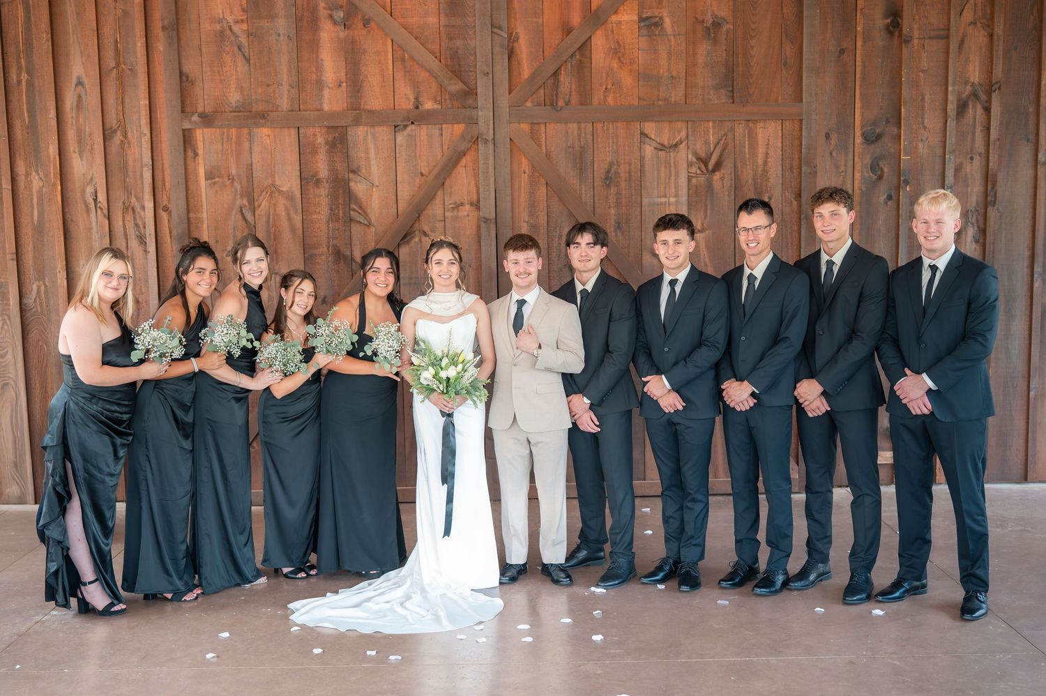 Wedding party poses in front of rustic barn doors, bridesmaids in green gowns and groomsmen in dark suits flanking the bride and groom.