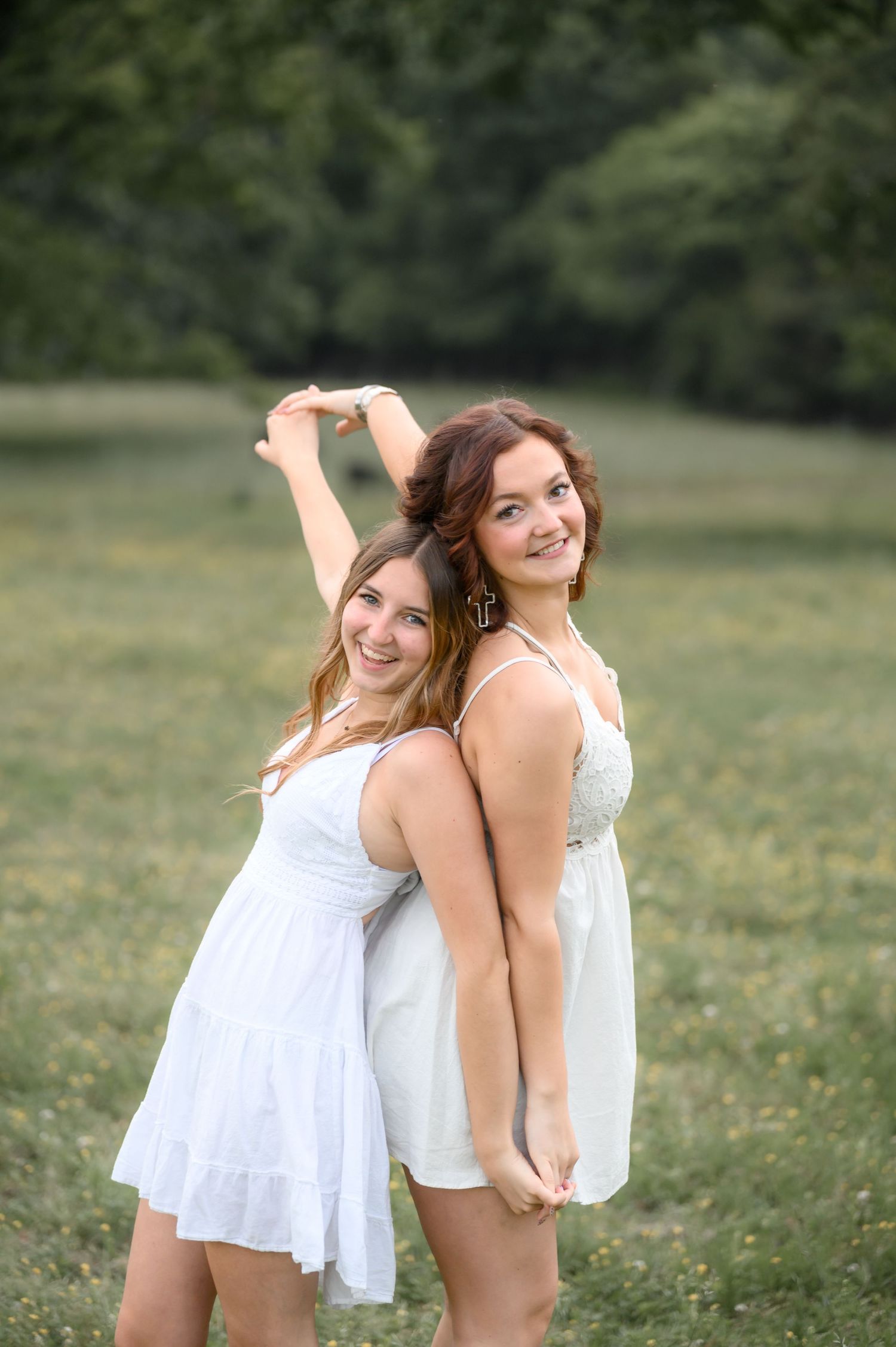 Two smiling young women in white dresses posing back-to-back in a green outdoor field.