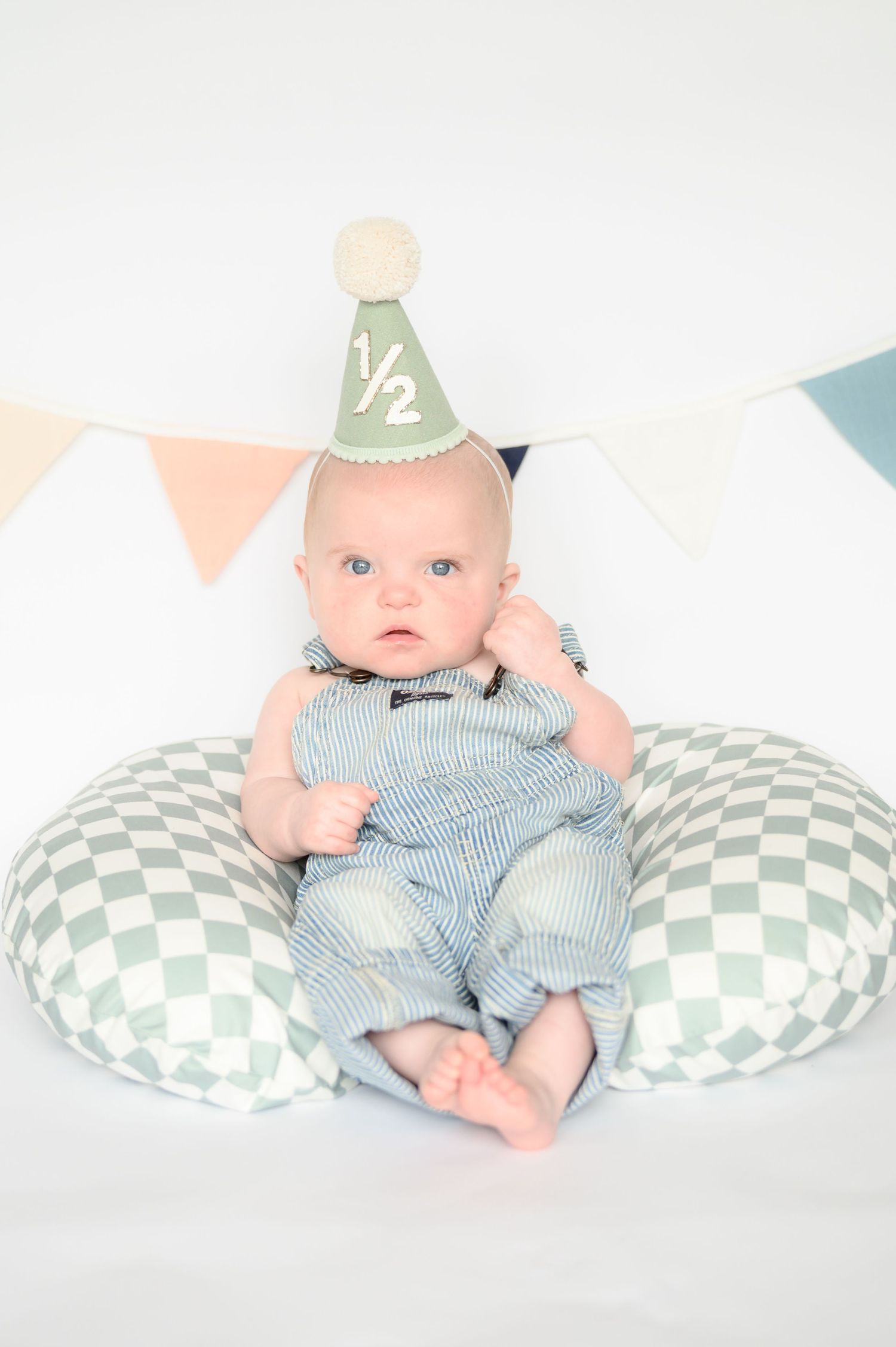Baby in striped OshKosh overalls wearing a green half birthday party hat sitting on checkered nursing pillow.
