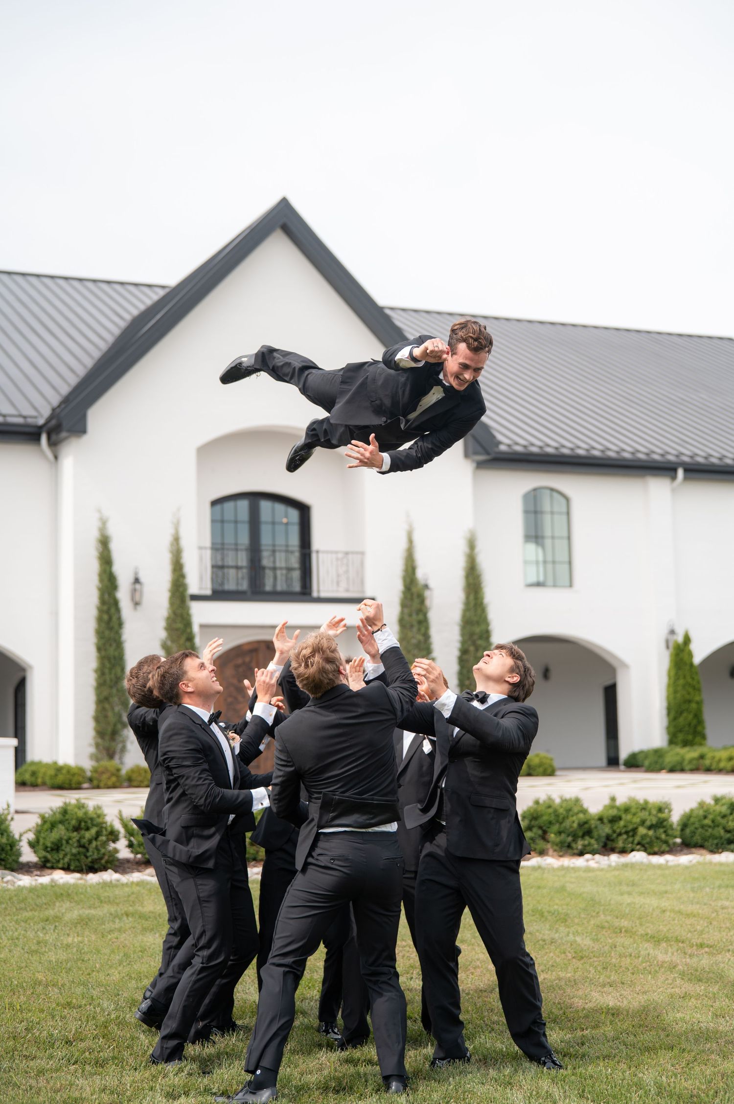 Groomsmen tossing groom into the air outside elegant white wedding venue.