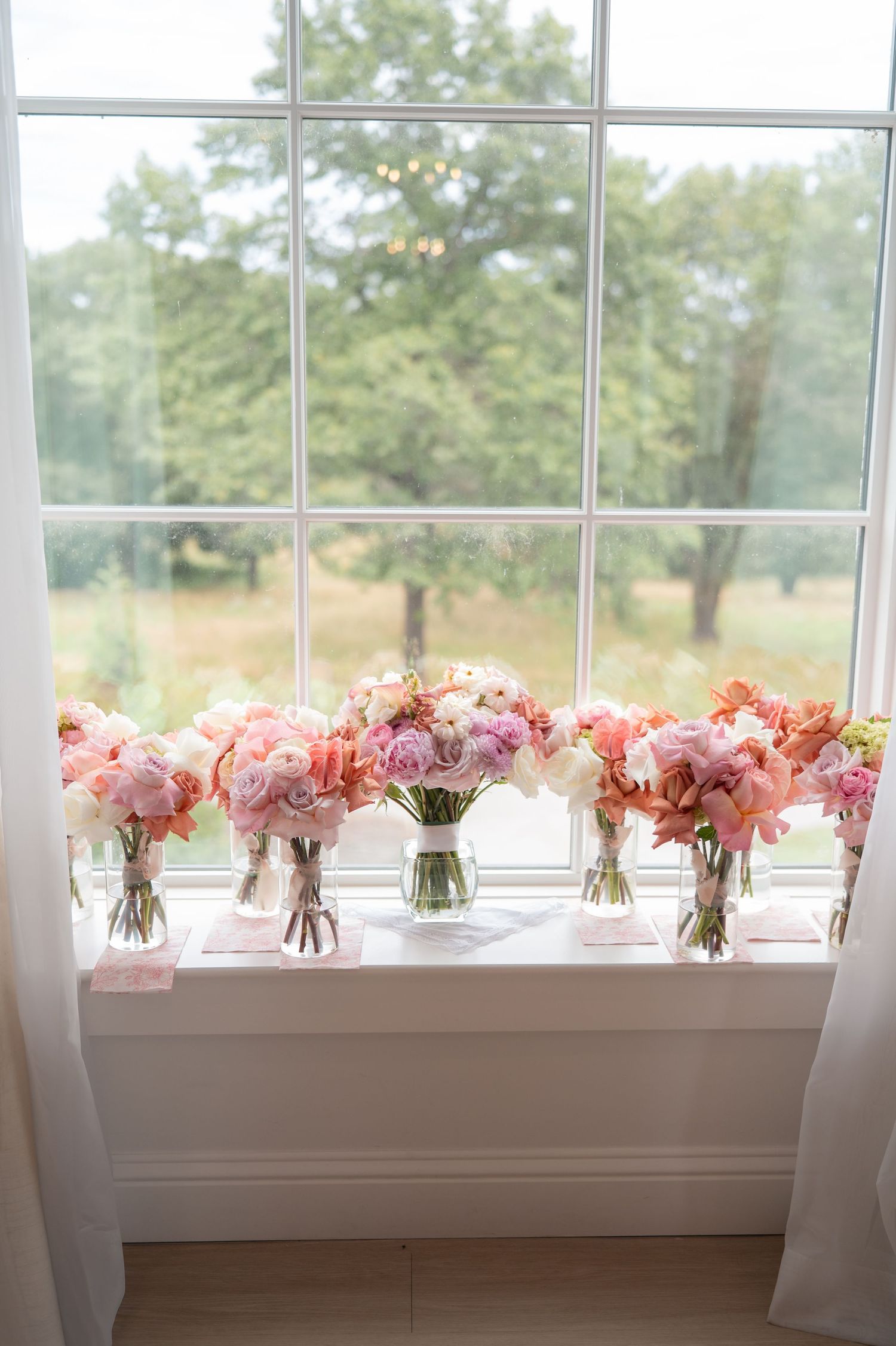 Row of pink and blush bridal bouquets displayed in glass vases along a bright window sill.