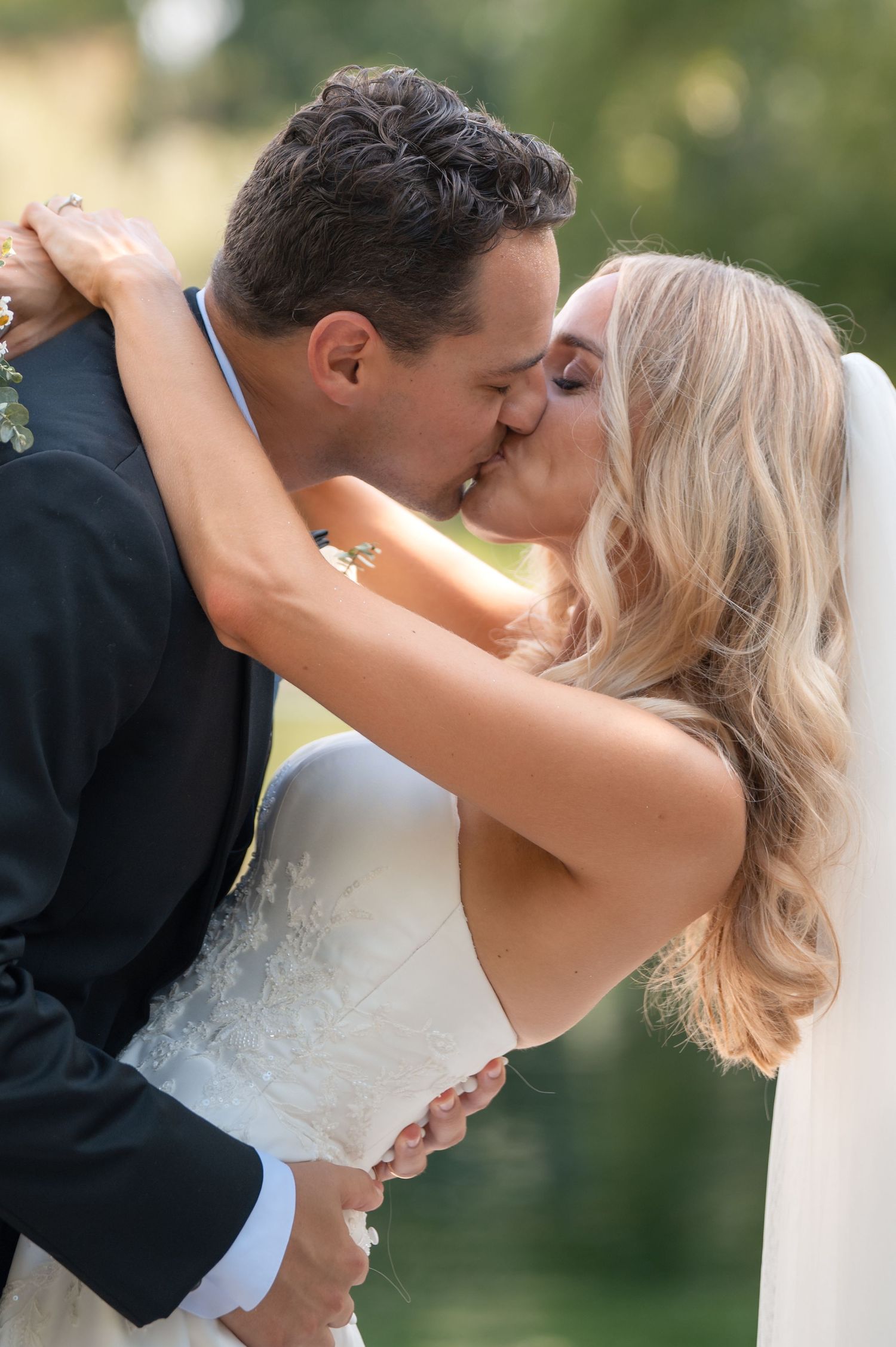 Bride and groom share passionate outdoor kiss, bride in strapless lace gown with veil.
