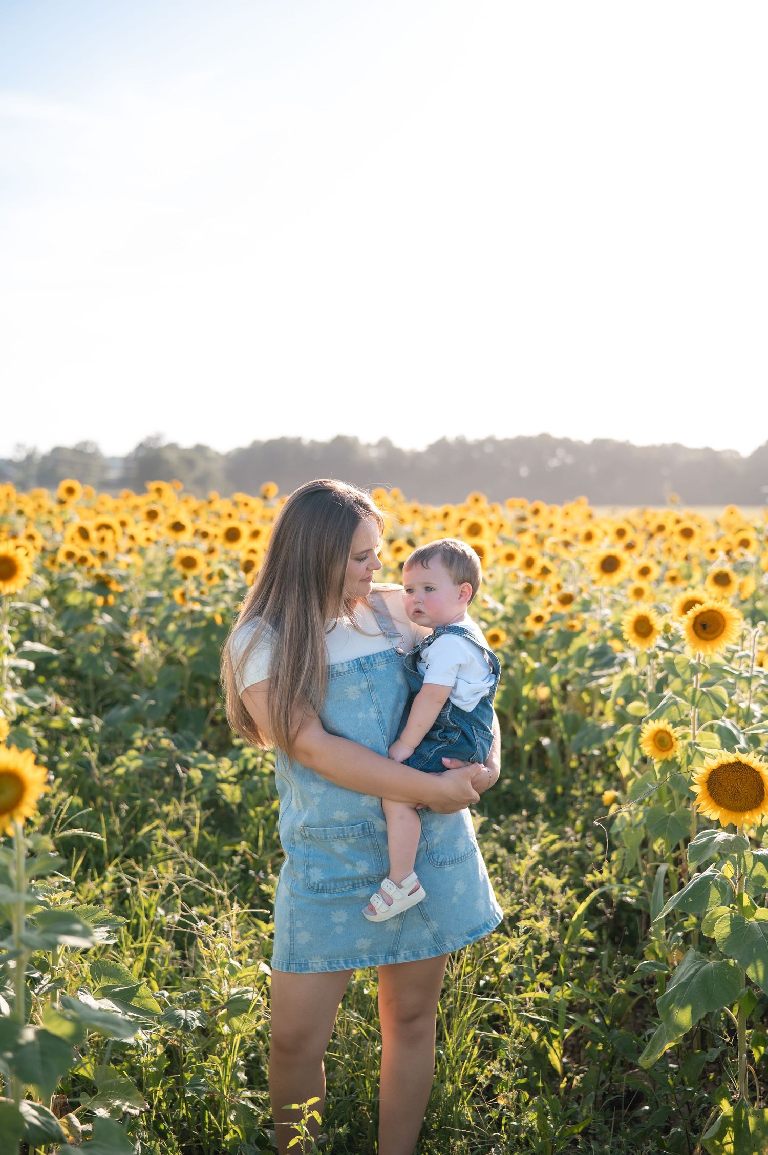 Mother in denim dress holding smiling baby in sunflower field with bright backlit sunshine.