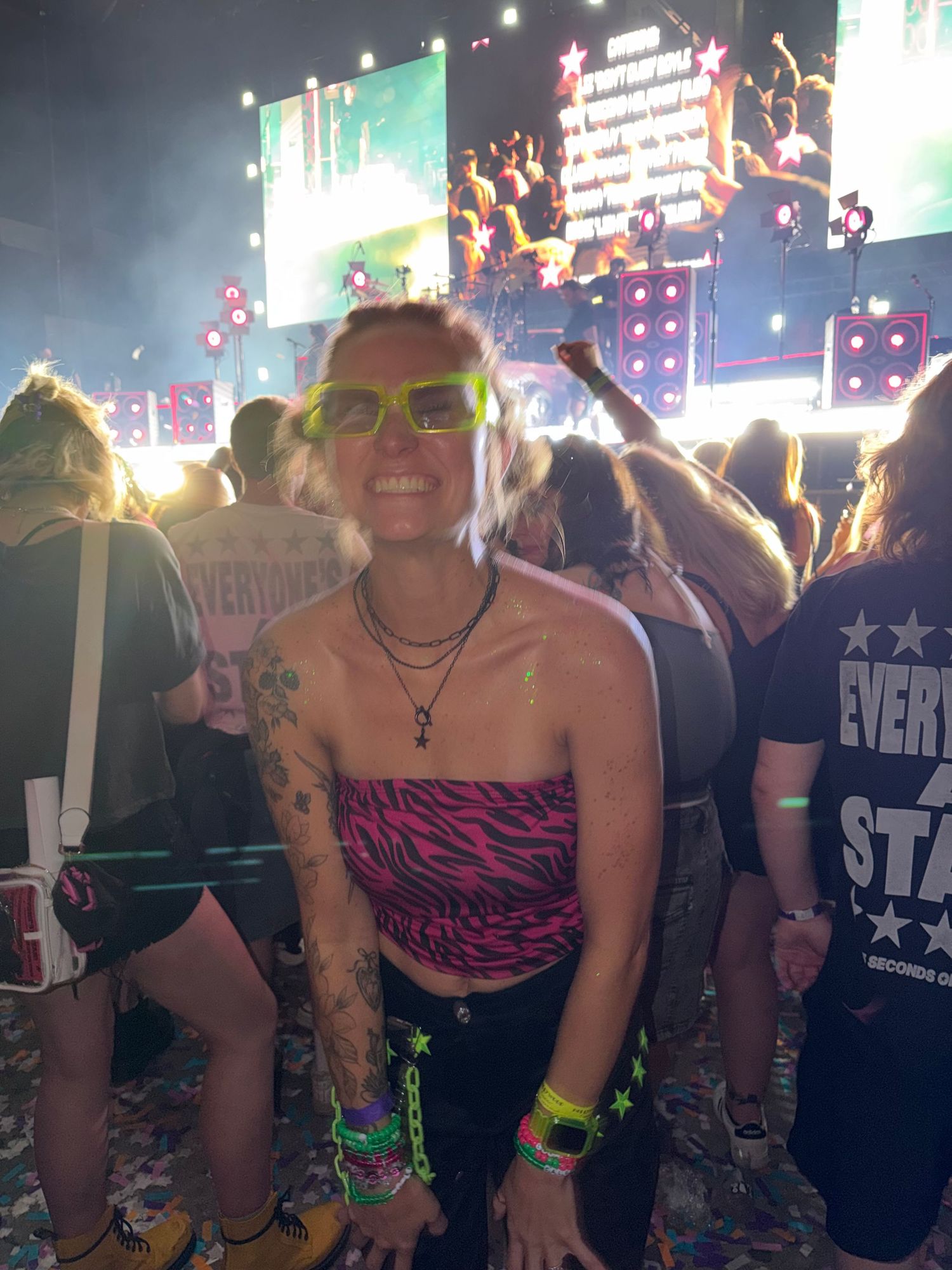 Woman in pink zebra tube top and green sunglasses smiles at a colorful music festival concert crowd.
