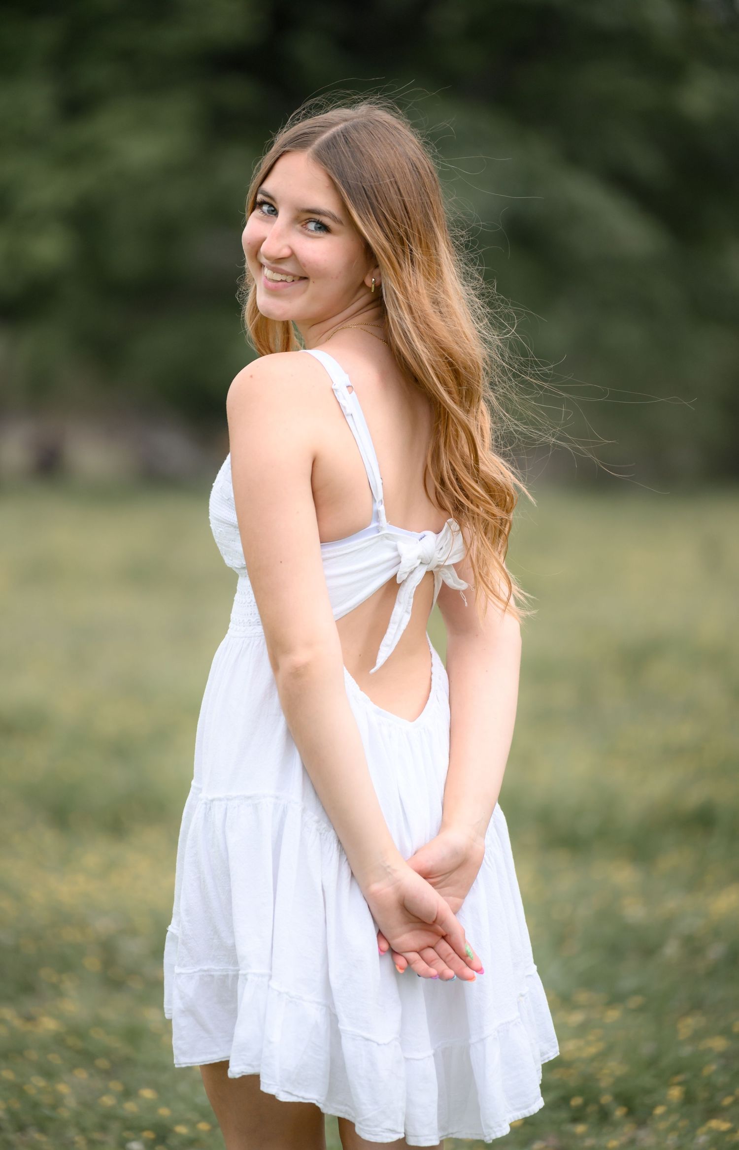 Young woman in white sundress smiling over her shoulder while standing in a green outdoor field.