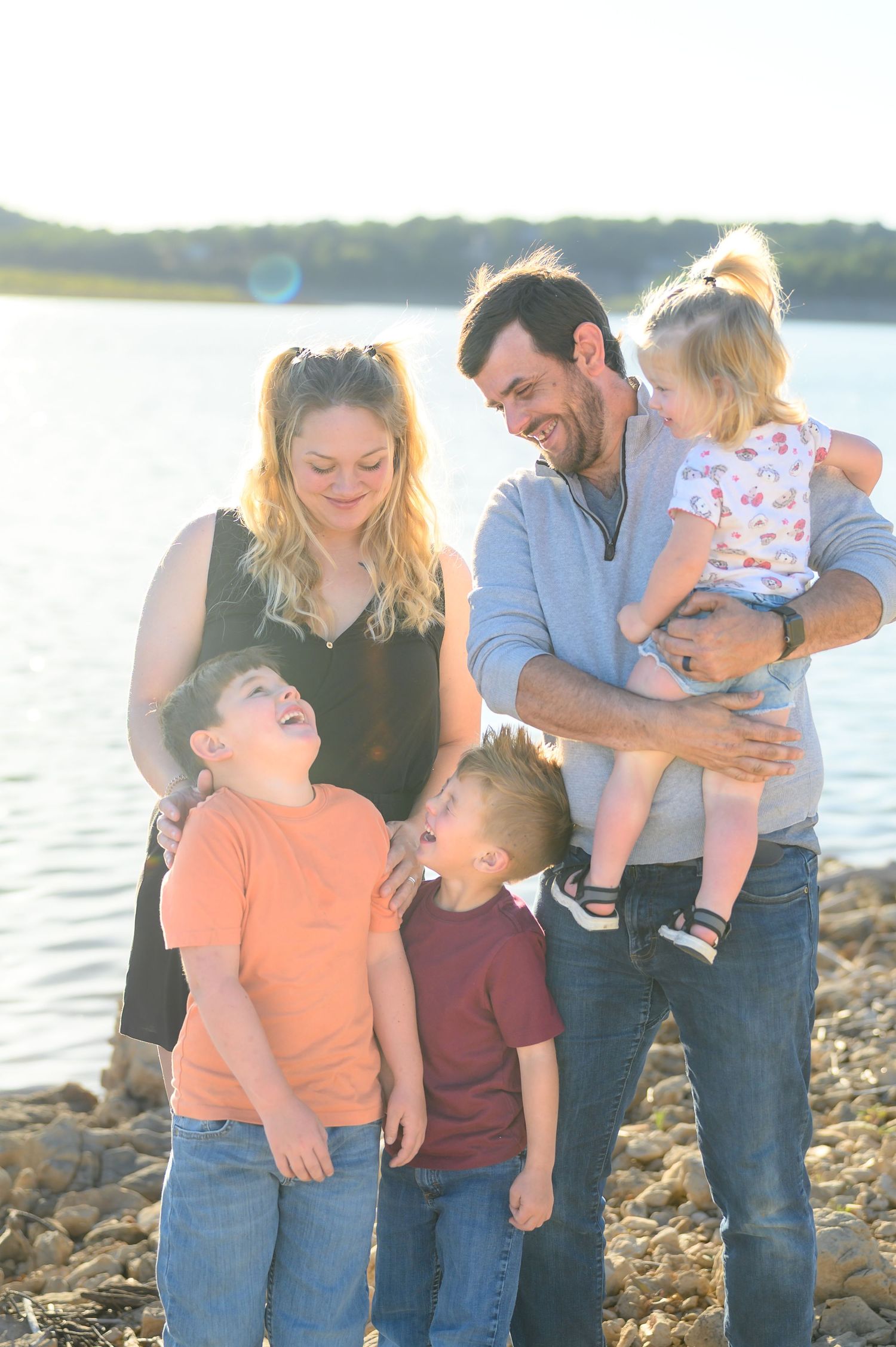 Happy family of five laughing together on a rocky lakeshore in warm golden sunlight.