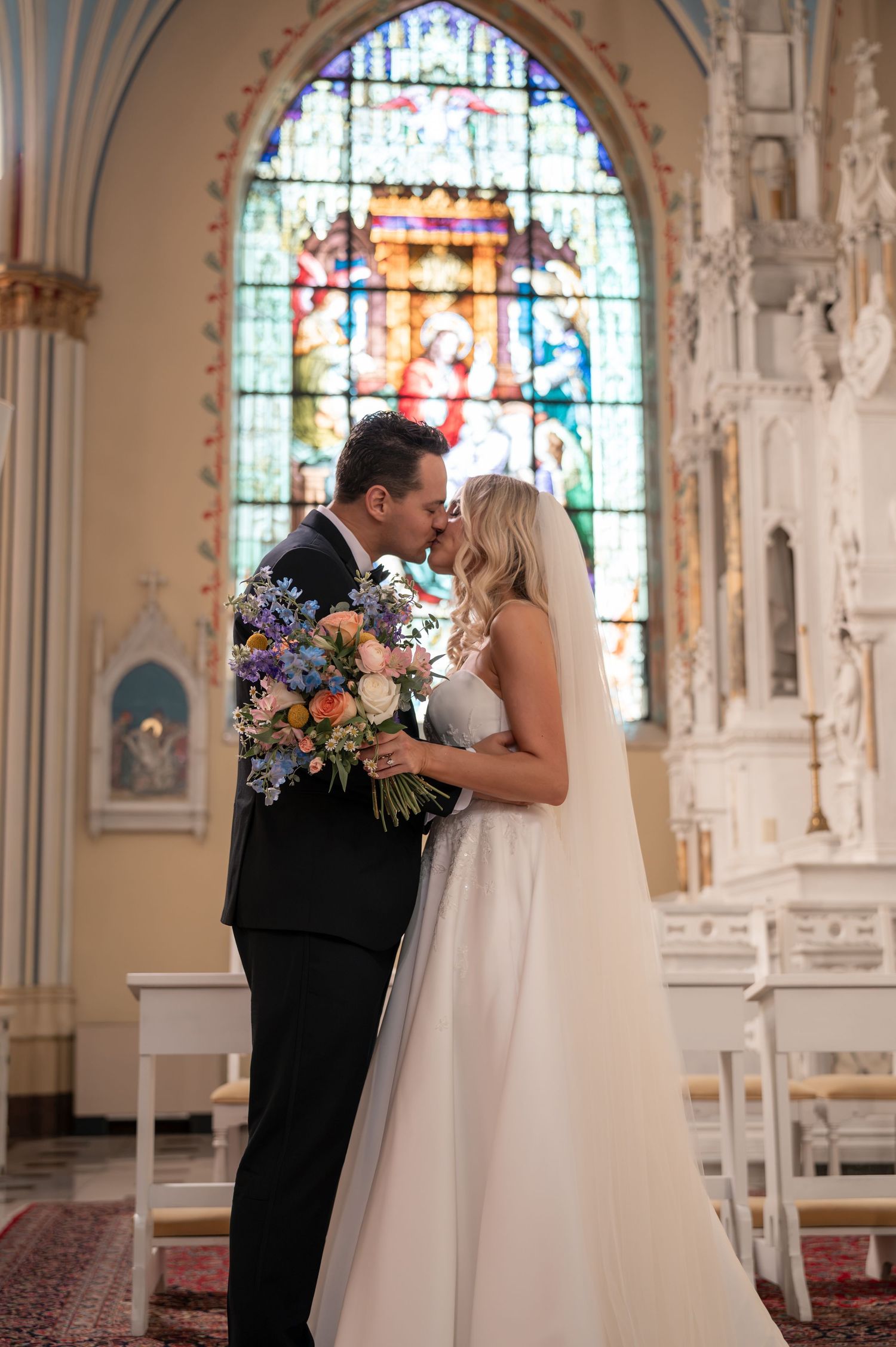 Bride and groom share kiss in ornate church with colorful stained glass windows.