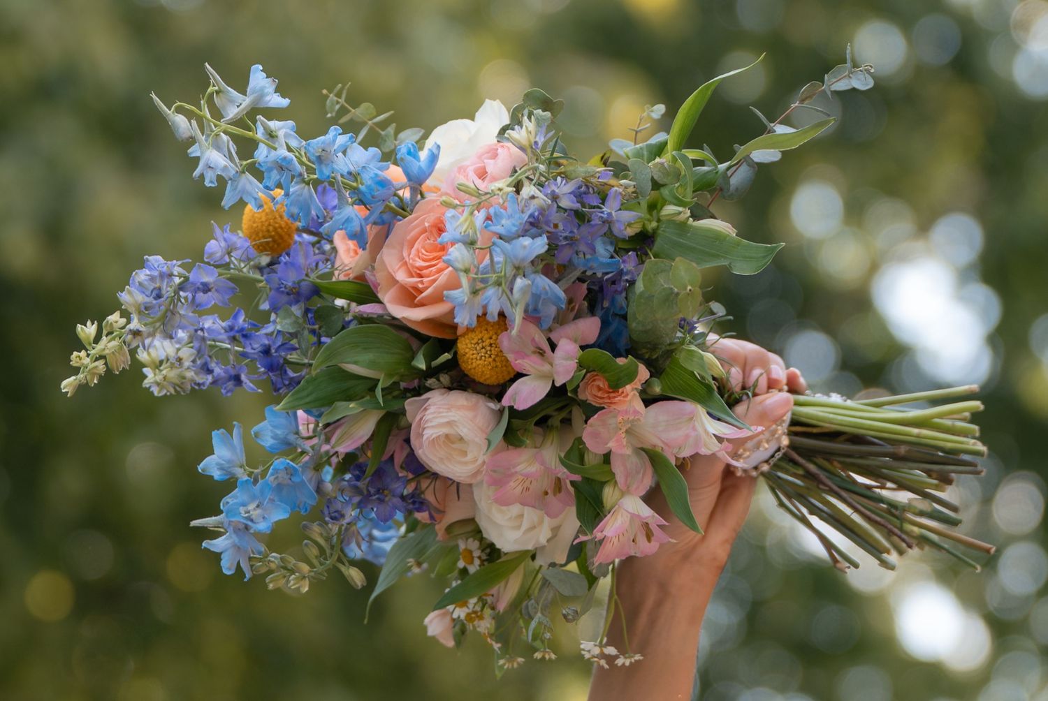 Hand holding a vibrant bridal bouquet with blue delphiniums, pink roses, and greenery outdoors.