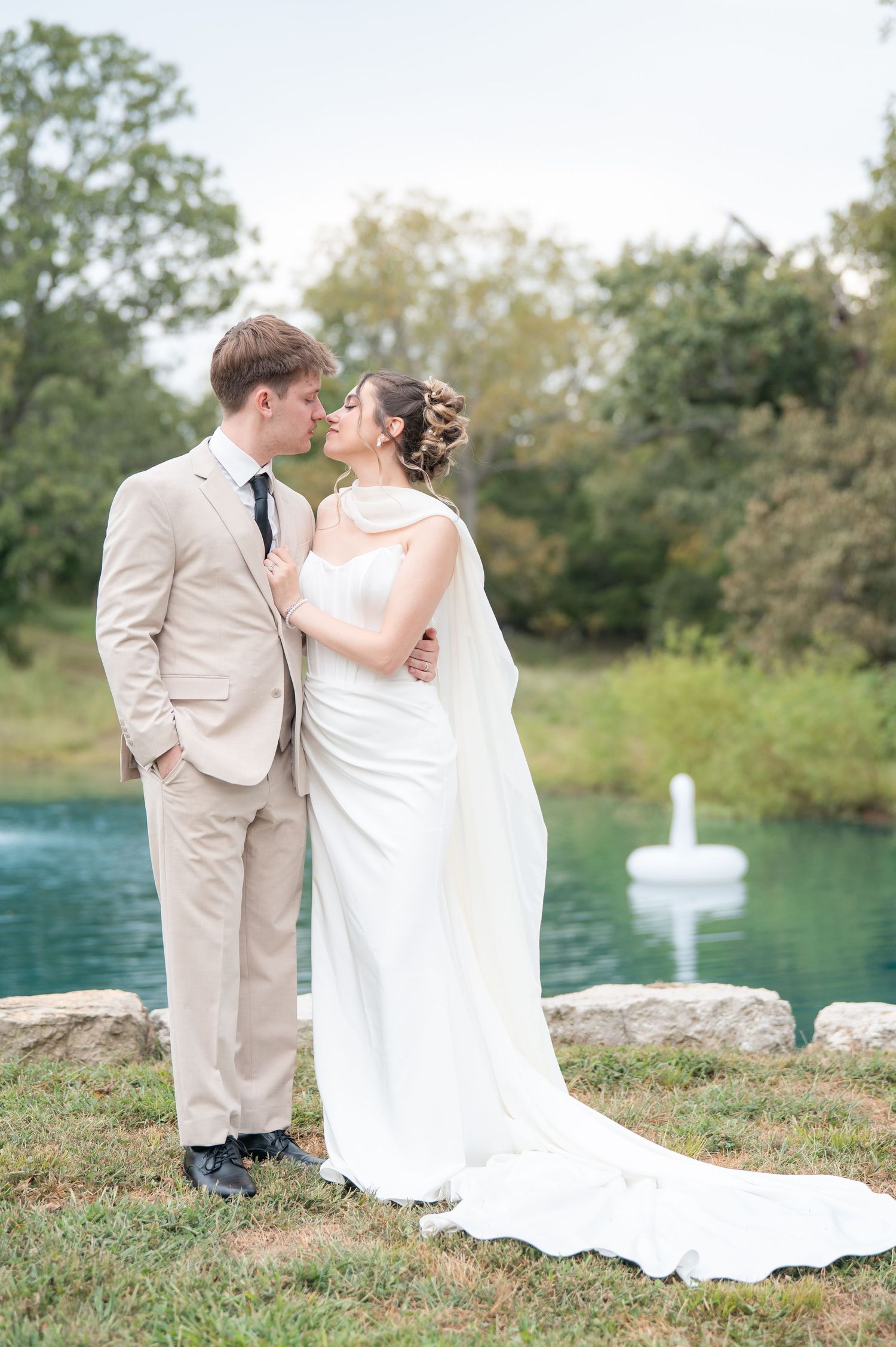 Bride and groom share a romantic moment by a serene blue pond, she in a white gown, he in a tan suit.